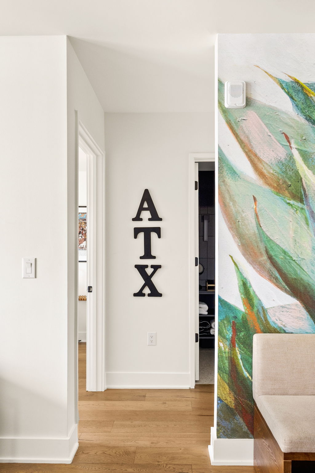 A glimpse into the hallway of a modern home, highlighting the light-colored hardwood flooring and crisp white walls. The space features a partial view of a vibrant mural, a built-in seating bench, and an inviting entryway into an adjacent room.