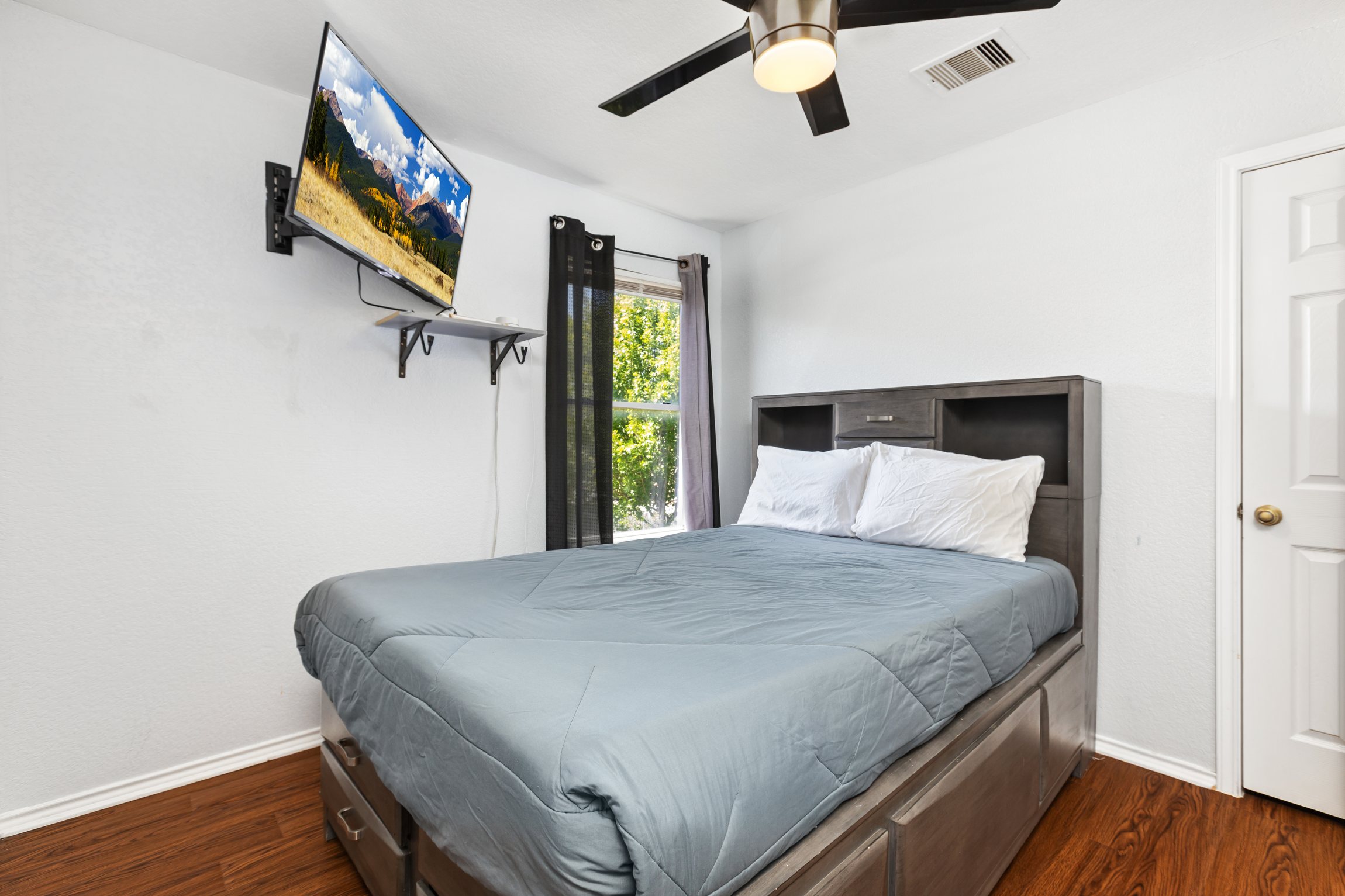 Queen bedroom with wall-mounted Smart TV, streaming, ceiling fan, hotel-quality bedding, and clothing storage. Room-darkening curtains and hardwood floors. One of 5 private bedrooms, Northeast San Antonio.