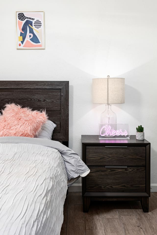A glowing "Cheers" neon sign, elegant bedside lamp, and soft pink pillow create an inviting boutique-hotel feel.