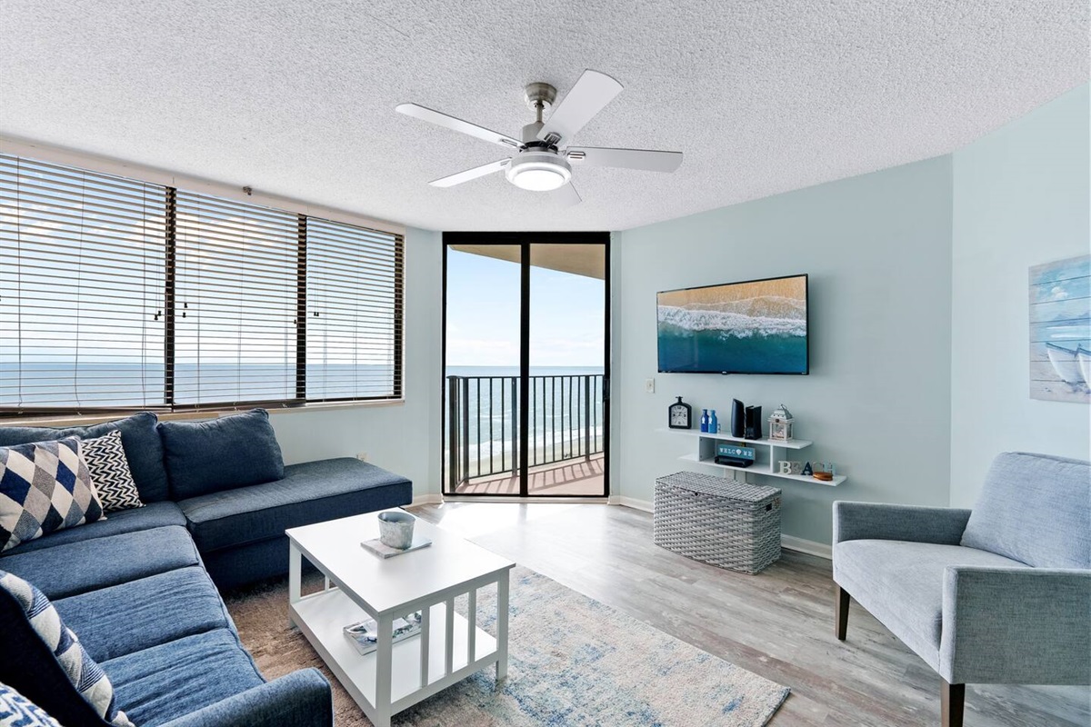 A chic TV lounge with flat tv screen , adaptable white and blue furnishings, large windows that invite abundant natural light, and a balcony showcasing stunning coastal views—ideal for unwinding and entertaining.
