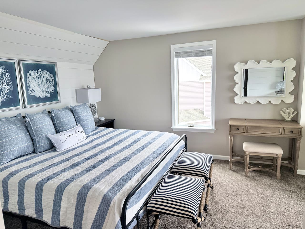 This bedroom features a cozy bed with blue and white striped bedding. The space is accented by a vanity with a unique, textured mirror and a matching stool, completing the tranquil, coastal-inspired design.