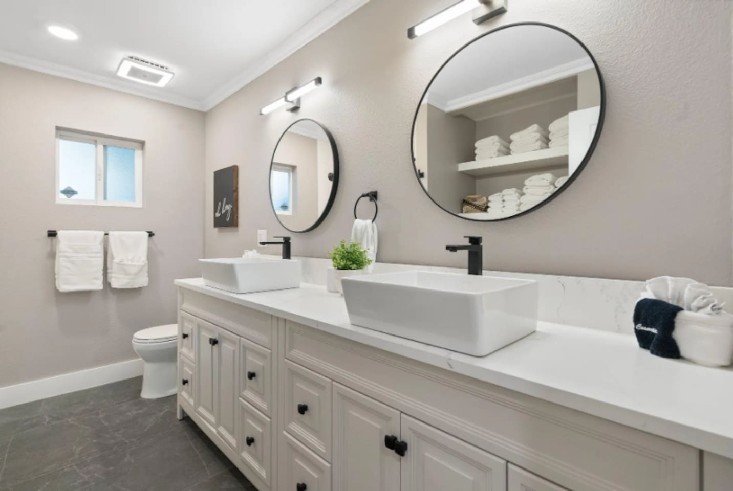 A long dual vanity with vessel sinks gives this bathroom plenty of room to spread out, while round mirrors and stacked fresh towels add a spa-like finish. Natural light from the window keeps the space feeling bright throughout the day.
