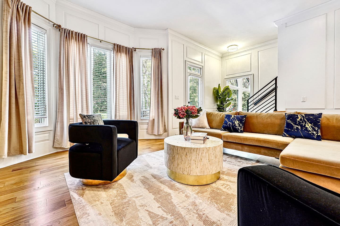 Situated next to the kitchen, this main-floor living room offers a warm, upscale ambiance with a brown L-shaped sectional, two black accent chairs, gold and black design accents, a smart TV, and a fireplace, perfect for entertaining or unwinding.