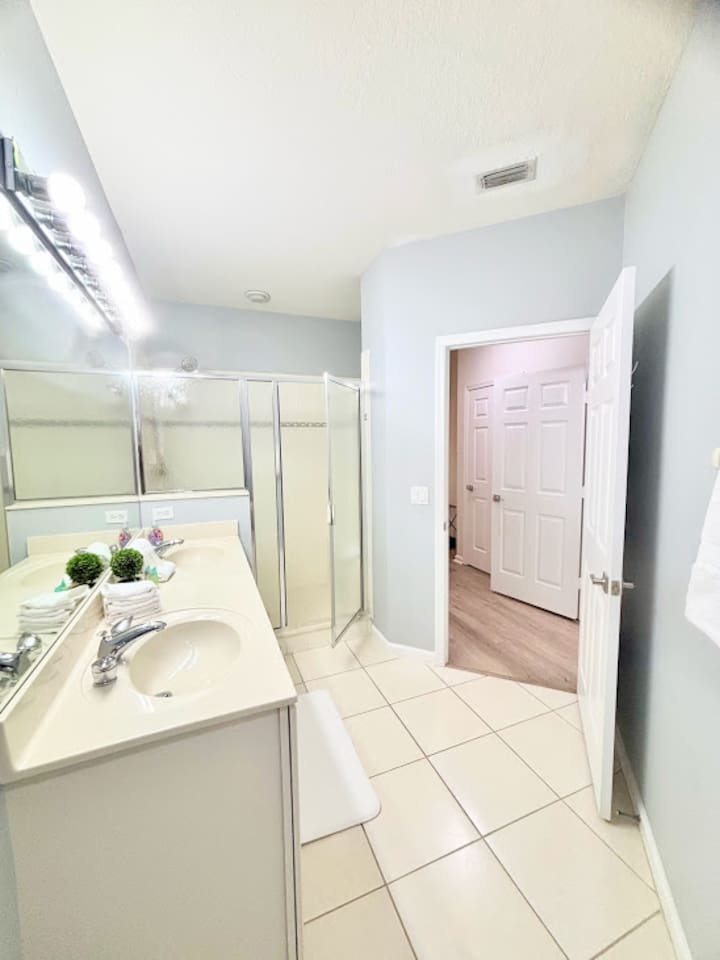 Bright bathroom with a spacious vanity, large mirror, tile flooring, and a walk-in shower with glass doors.