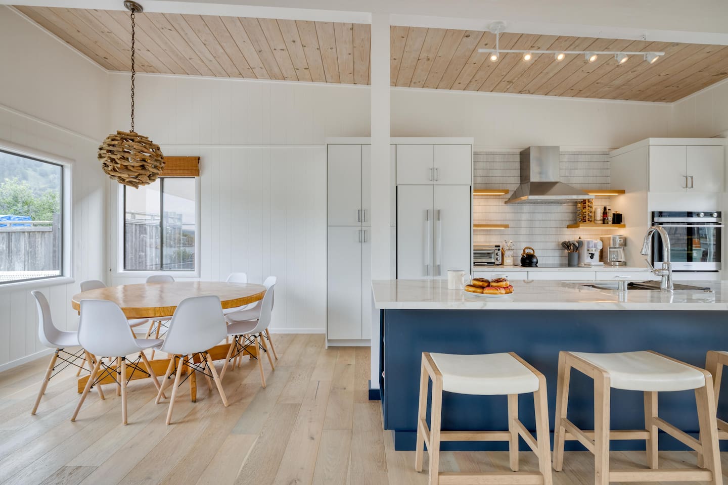 - Dining space with round table and modern chairs for shared meals  
- Kitchen features a navy island with bar seating for casual dining  
- Light wood flooring and ceiling create a warm, coastal inspired vibe  

