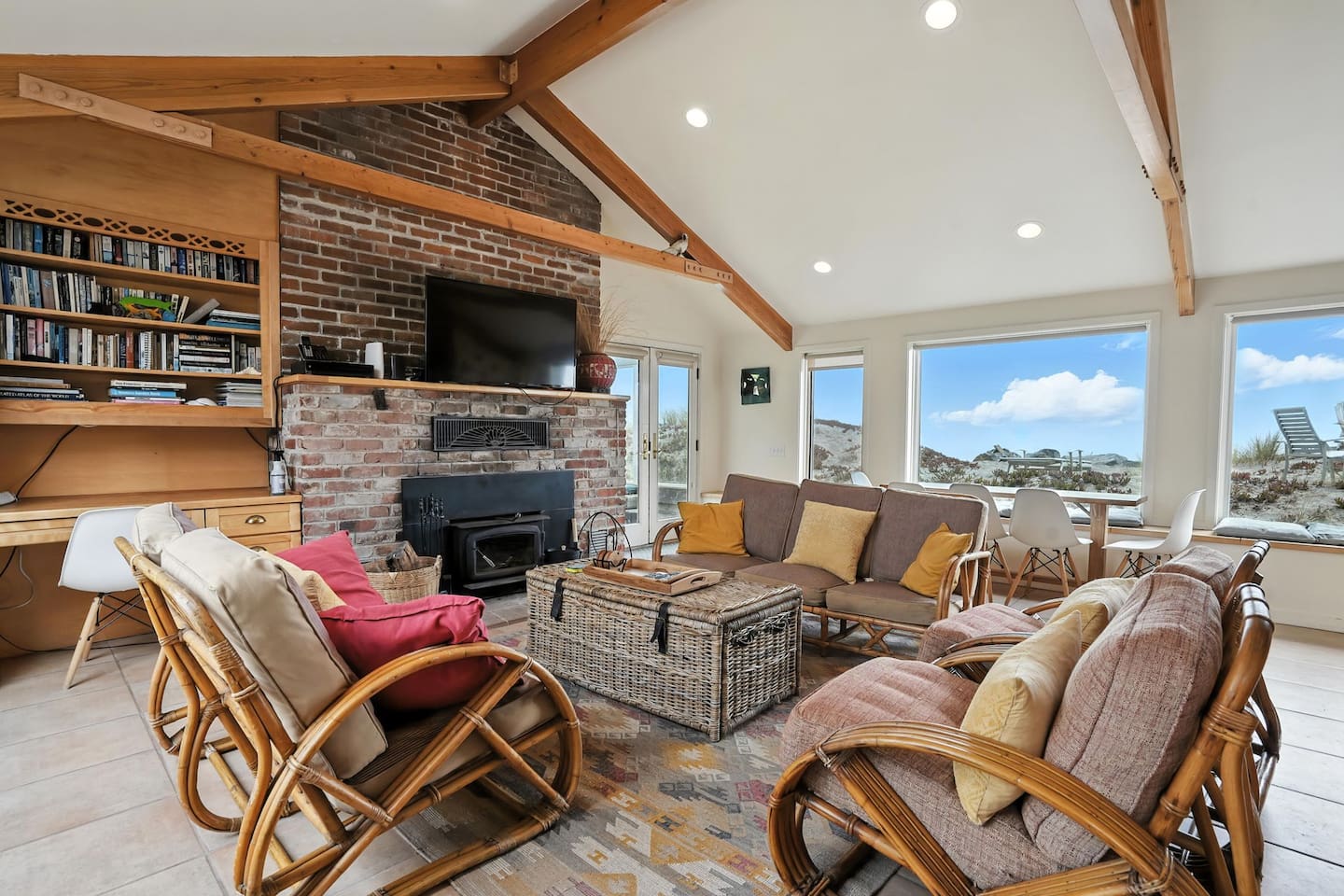 - Cozy living room with vaulted ceilings, rattan furniture, and vibrant pillows  
- Brick fireplace with mounted TV for a warm and inviting atmosphere  
- Large windows offer sweeping views of the dunes and ocean beyond  
