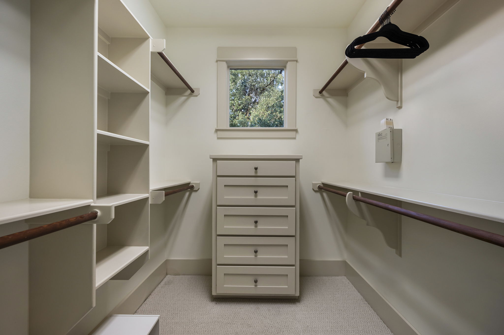 A bright and organized walk-in closet featuring built-in shelving, hanging rods, and a central dresser with drawers, illuminated by a window.