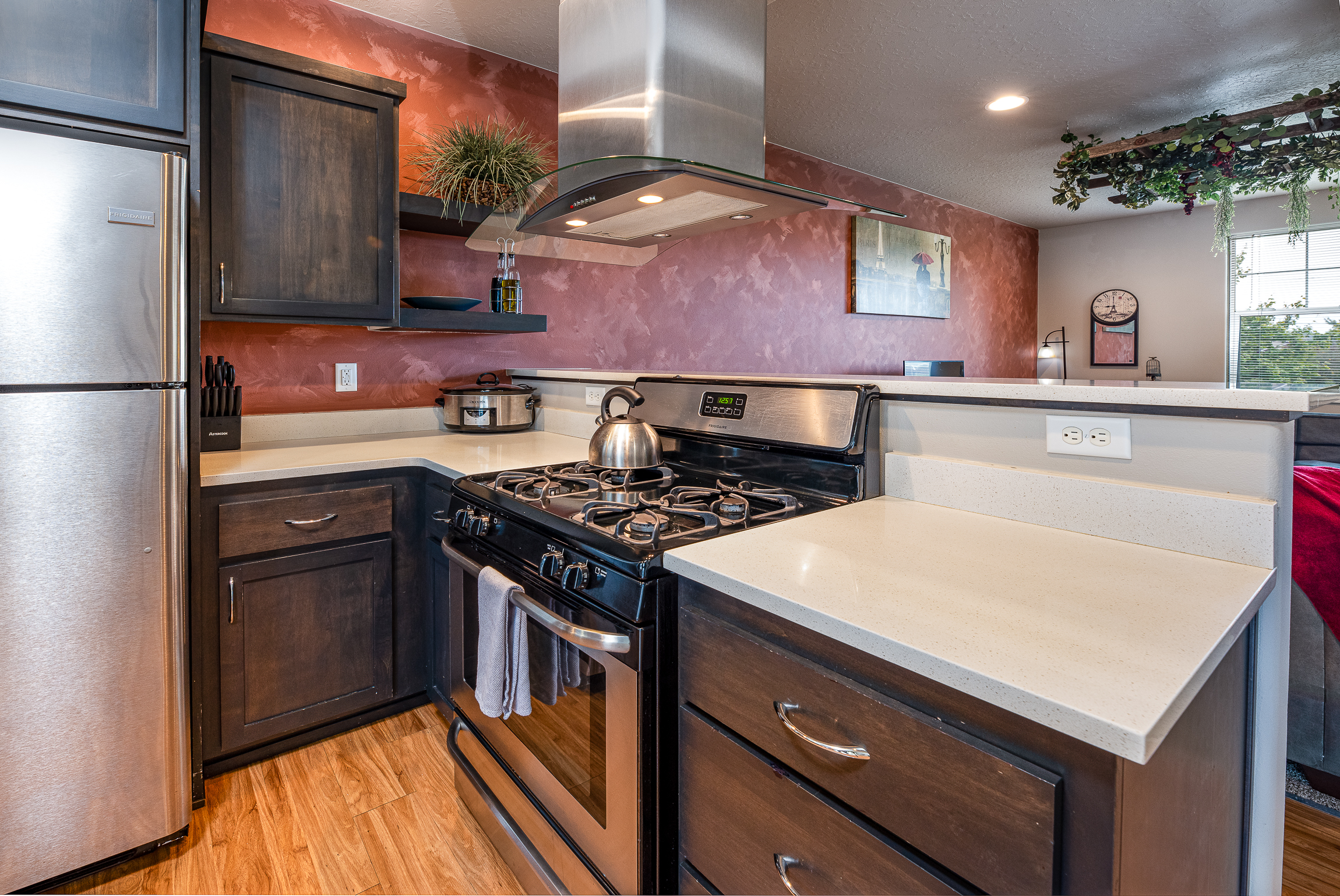 A modern kitchen featuring a stainless steel fridge, black gas stove with a kettle, wooden cabinetry, a light countertop, and decorative plants, set against a textured reddish wall with a wall clock and a framed picture.