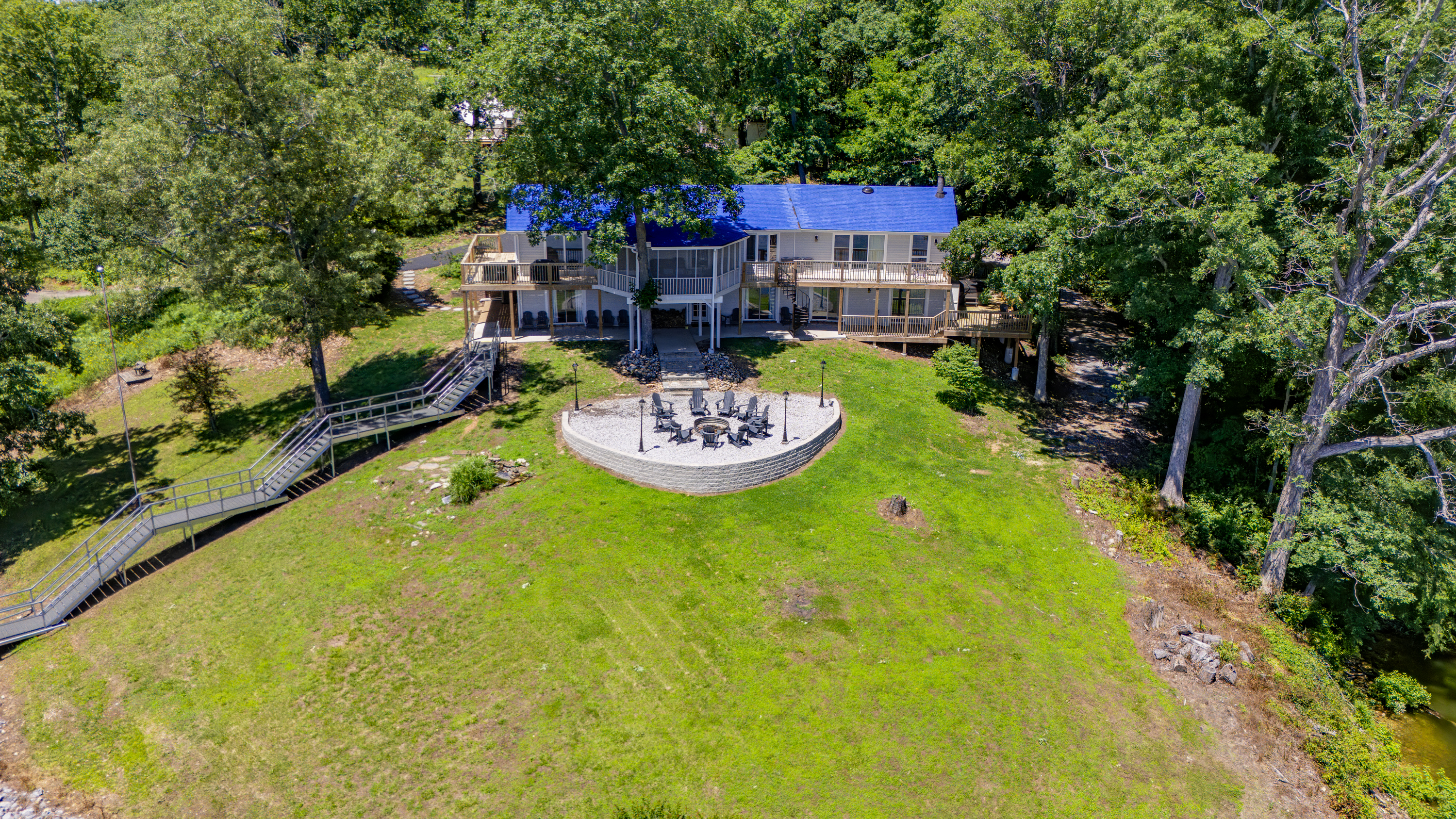A close aerial highlights the home, spacious lawn, circular firepit area, and walkway toward the private dock.