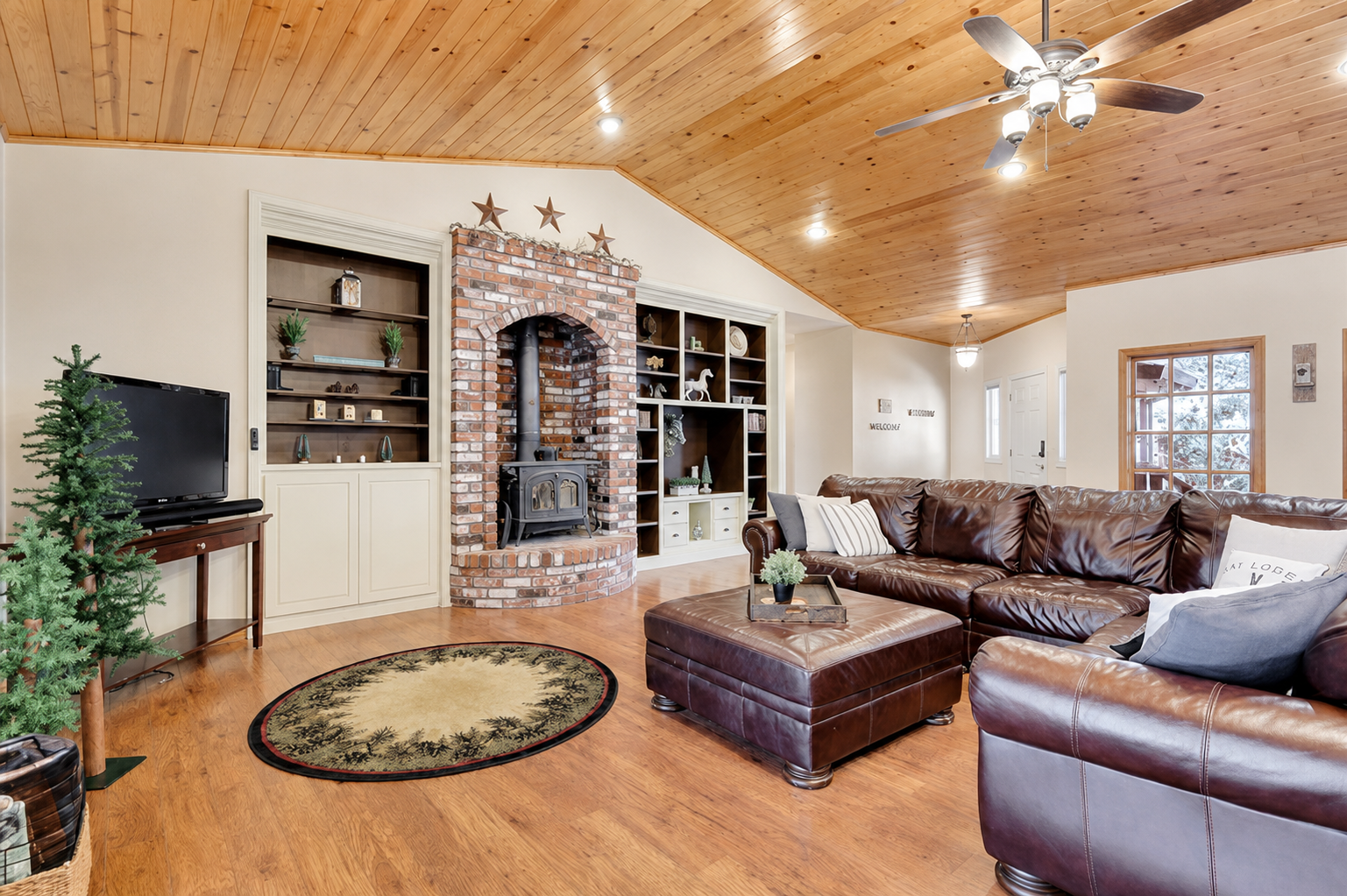 Living room with vaulted wood ceiling, leather sectional seating, brick fireplace, built-in shelving, and TV for cozy cabin downtime.