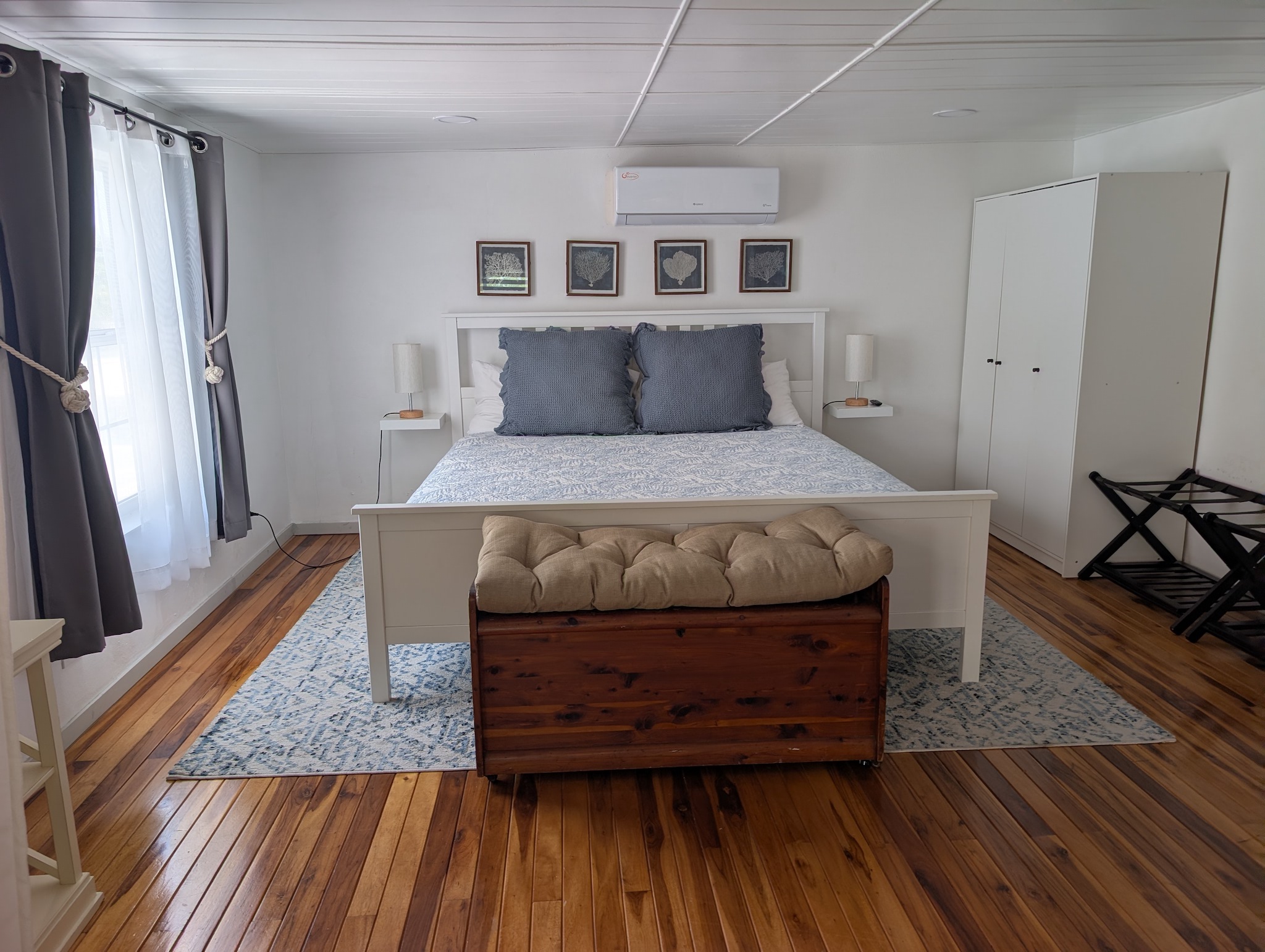 Upstairs master bedroom suite at Villa Coral featuring a white wooden king bed frame, blue coastal linens, rich hardwood flooring, a cedar chest bench, and a split-unit air conditioner in Dorado, Puerto Rico.

