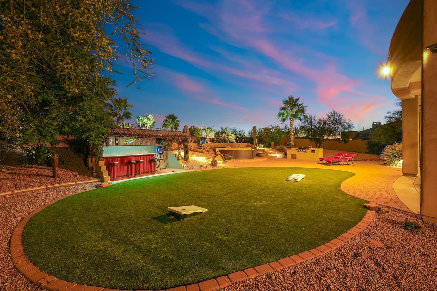 Backyard at dusk showcasing the turf lawn, cornhole setup, and vibrant outdoor lighting.