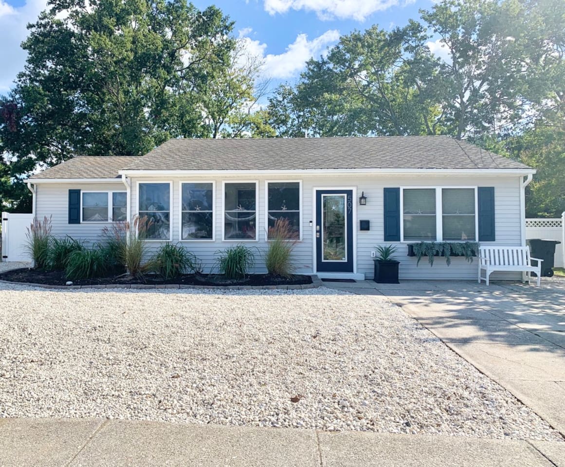 Enjoy the curb appeal of this charming cottage. The front features a glass-paneled navy door, large windows, and a cozy white bench. With a clean stone yard and lush greenery, it’s a welcoming first look at your coastal home.