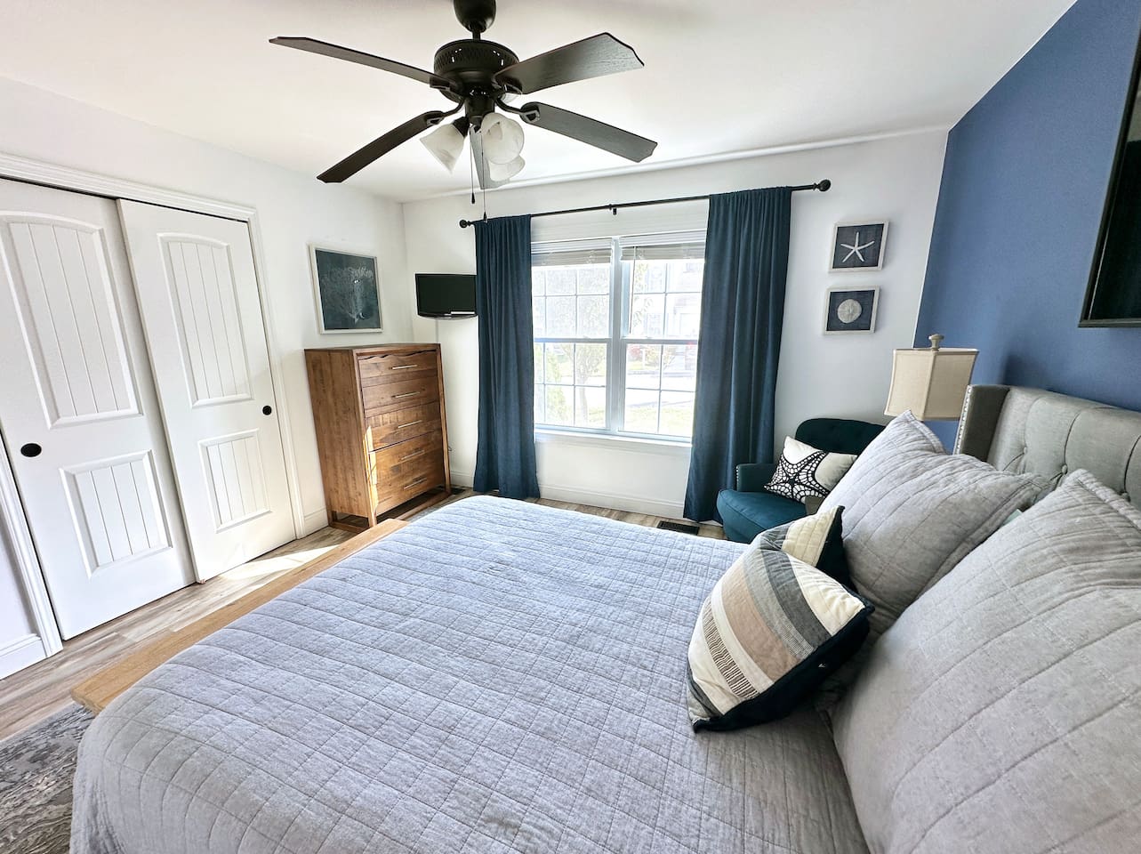 This bedroom offers a cozy layout with a large window. It features a tall wooden dresser, a double-door closet, and a comfortable bed. A blue accent wall and teal armchair add a stylish touch to the space.