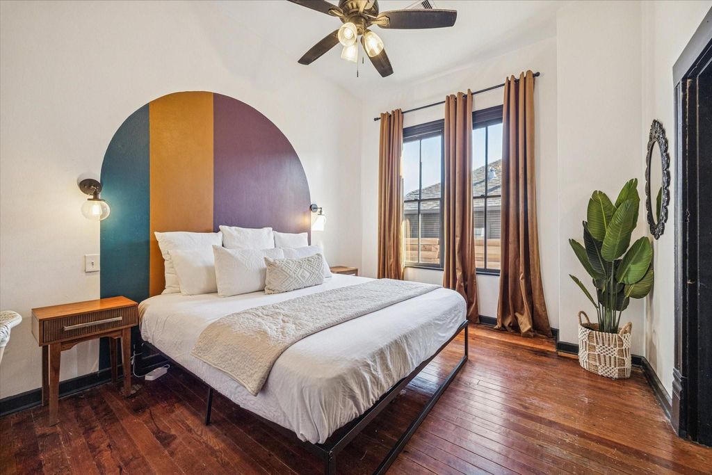 This bedroom makes a bold impression with original hardwood floors, tall windows wrapped in rich millwork, stylish lighting, and a striking accent wall with sconce lighting that adds personality without sacrificing warmth or comfort.