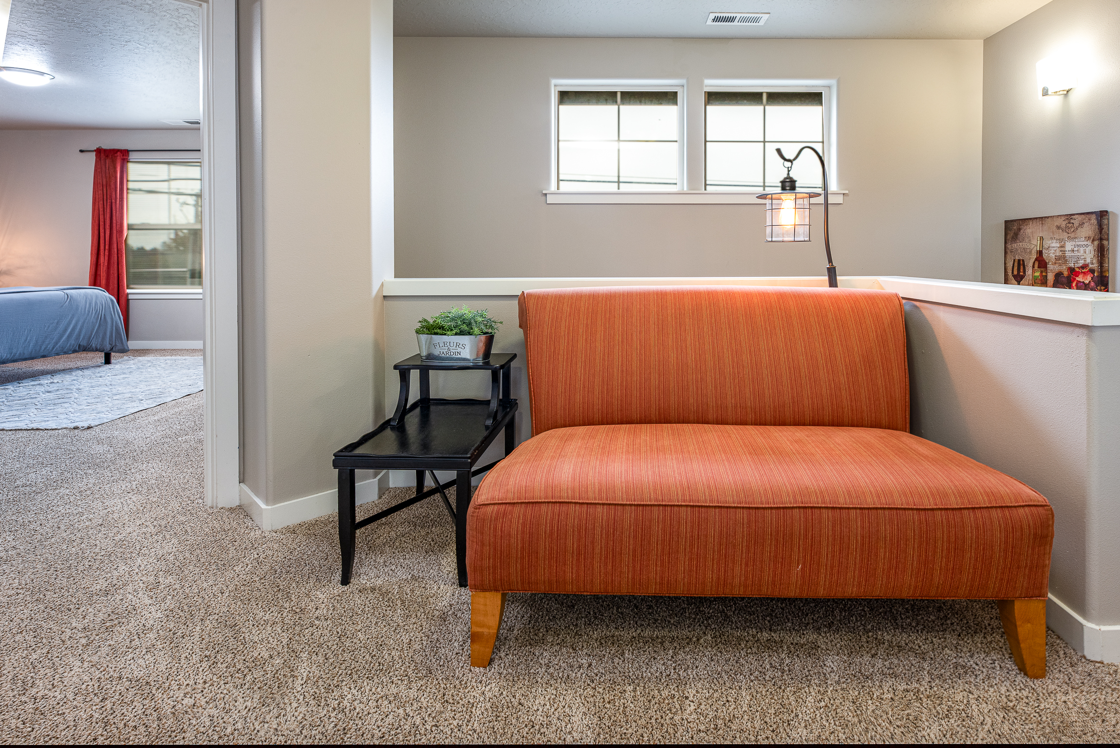 A cozy corner features an orange striped loveseat beside a black side table with a small potted plant, set in a well-lit room that opens to another space with a blue bedspread and red curtains in the background.