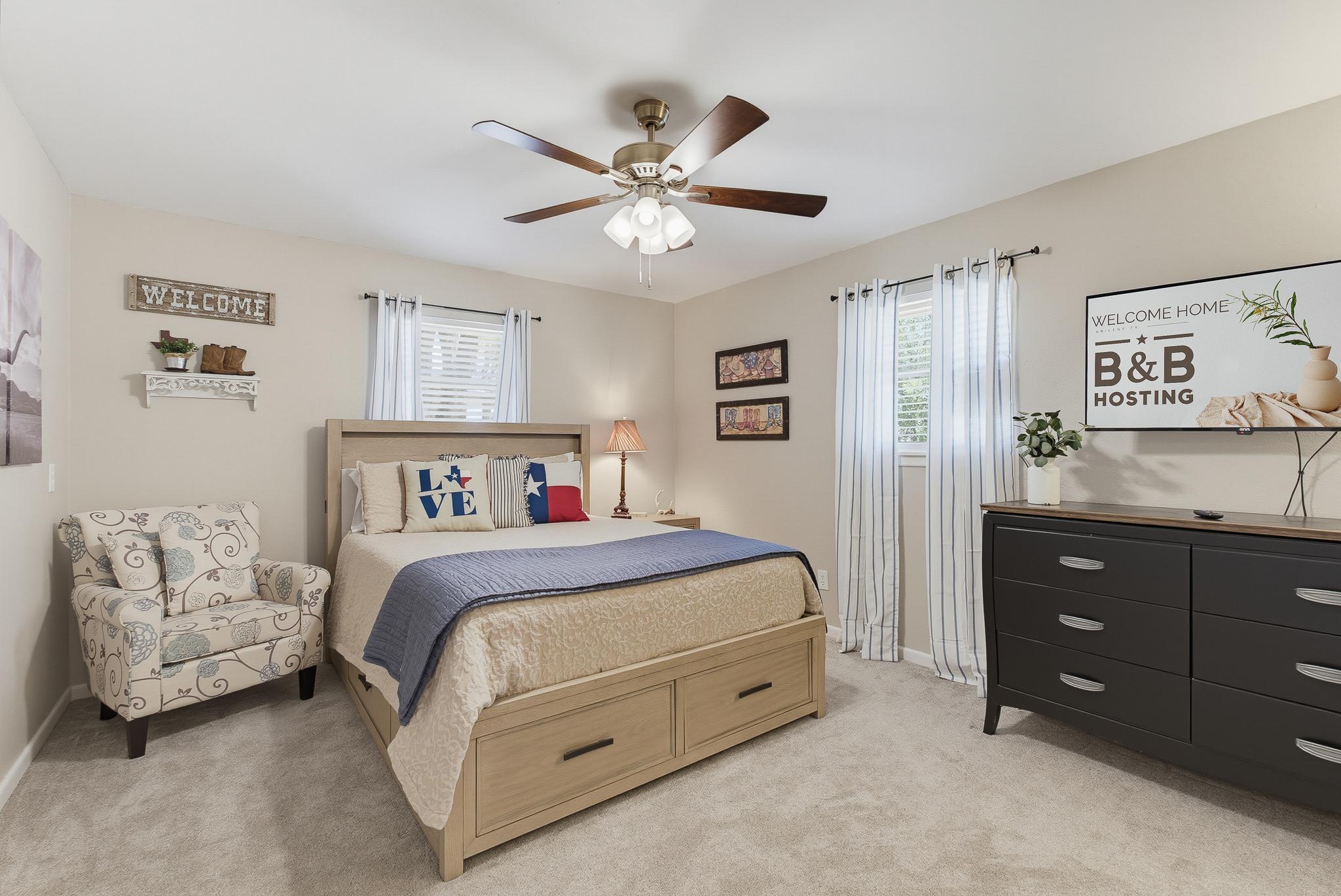 Texas-Themed Bedroom A warm, welcoming bedroom featuring a wood-toned storage bed dressed with "LOVE" and Texas-flag throw pillows, a cozy floral accent chair, wall-mounted shelf with rustic "Welcome" sign, and a large dark dresser with wall-mounted TV. 