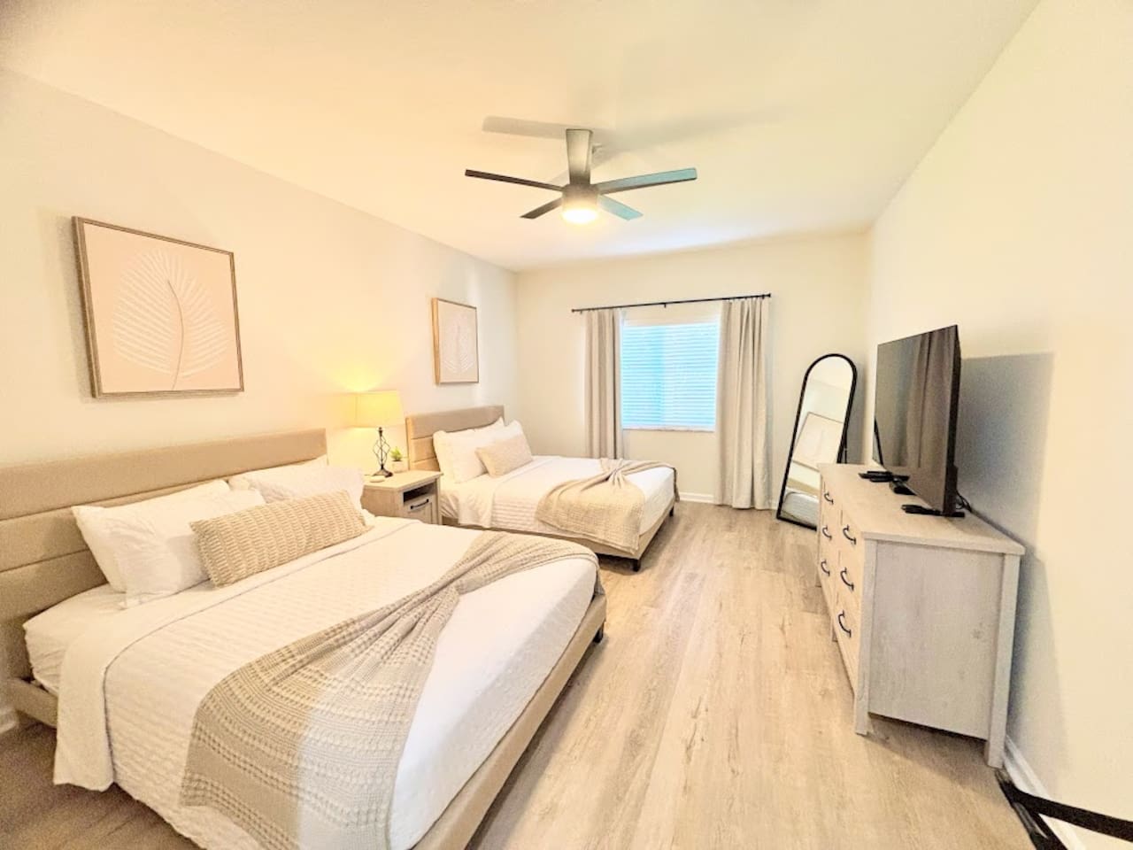 Bright and modern bedroom featuring two comfortable beds, wood-look flooring, ceiling fan, dresser storage, and a TV for a relaxing stay.
