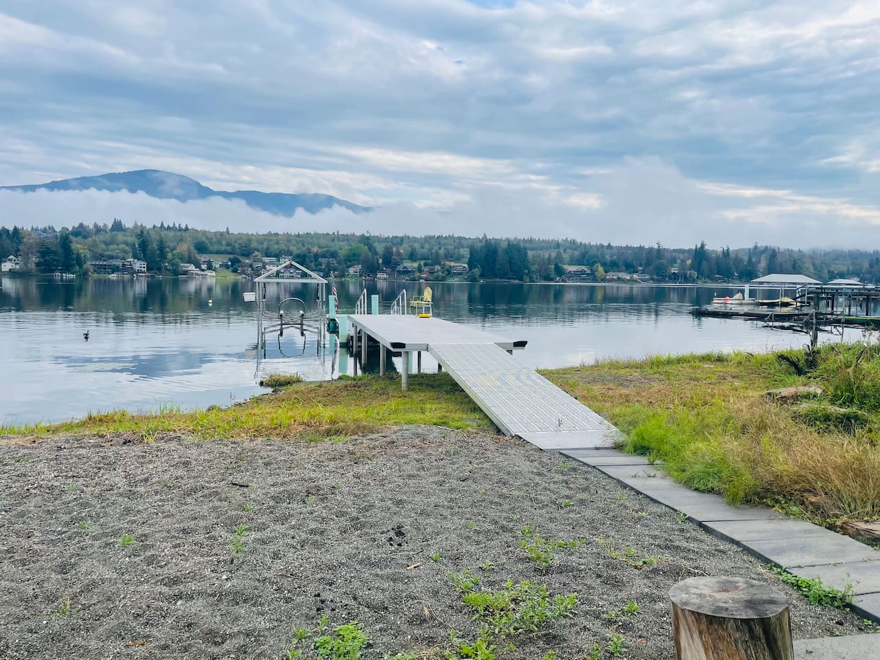 A dock stretches into the calm lake, accessible by a pathway with sand on one side and grass on the other. It’s perfect for mooring a boat or sitting by the water, blending natural textures with the peaceful lakeside setting.