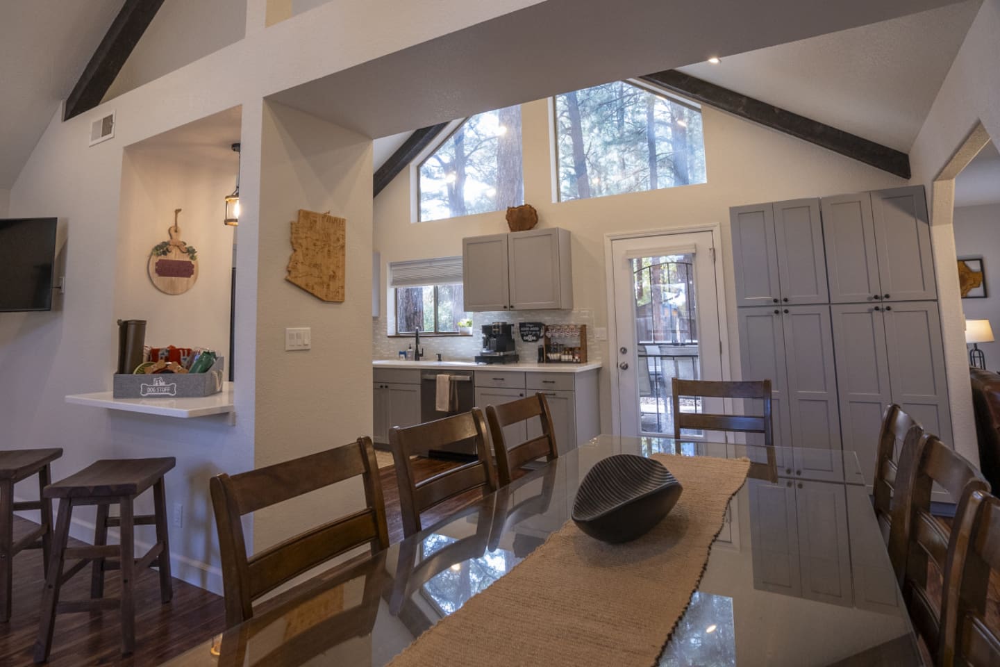 Dining space with a large table, nearby kitchen, and bright windows overlooking the trees.