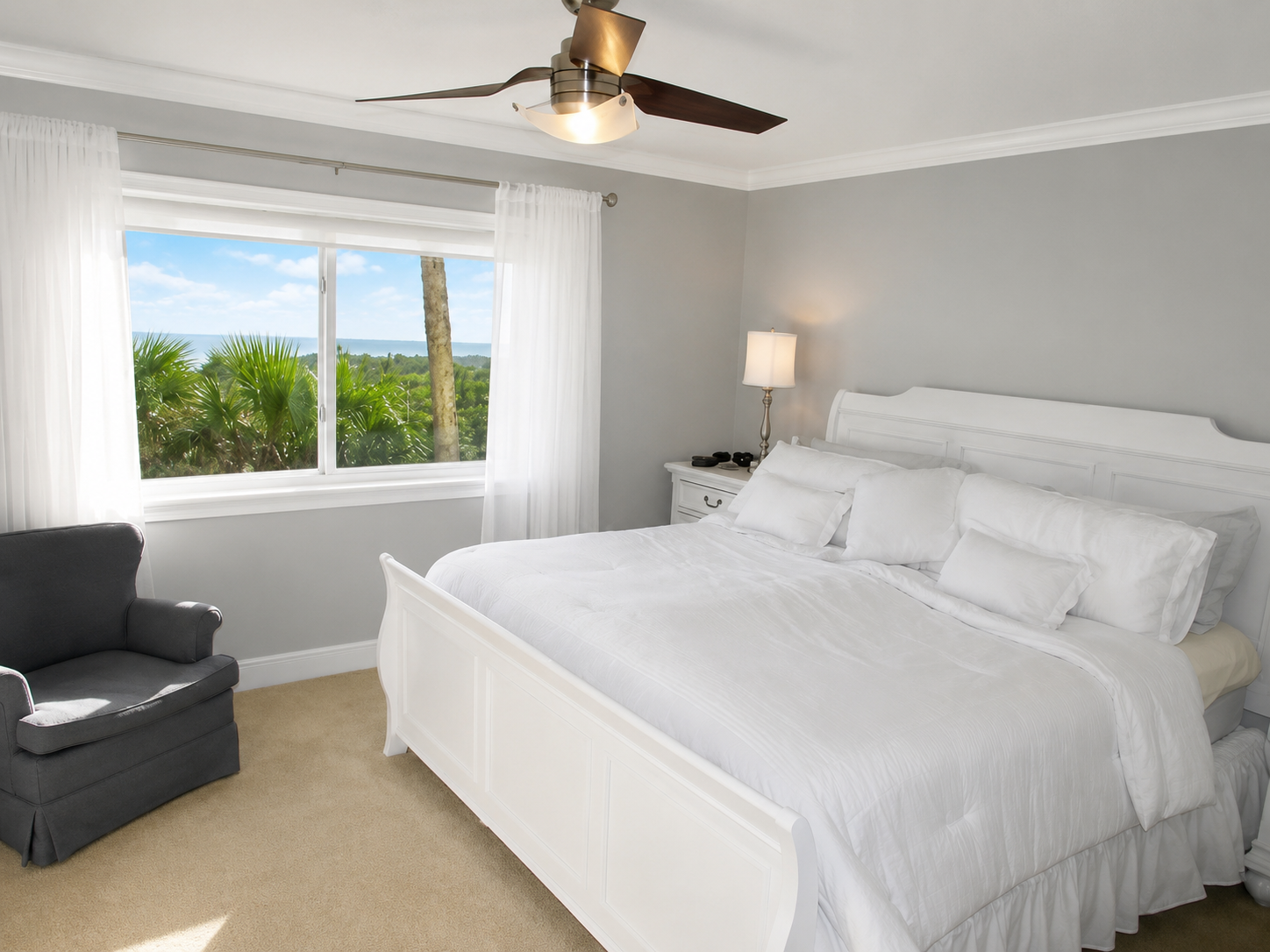 Bright bedroom with white bedding, gray armchair, and serene palm tree view outside.
