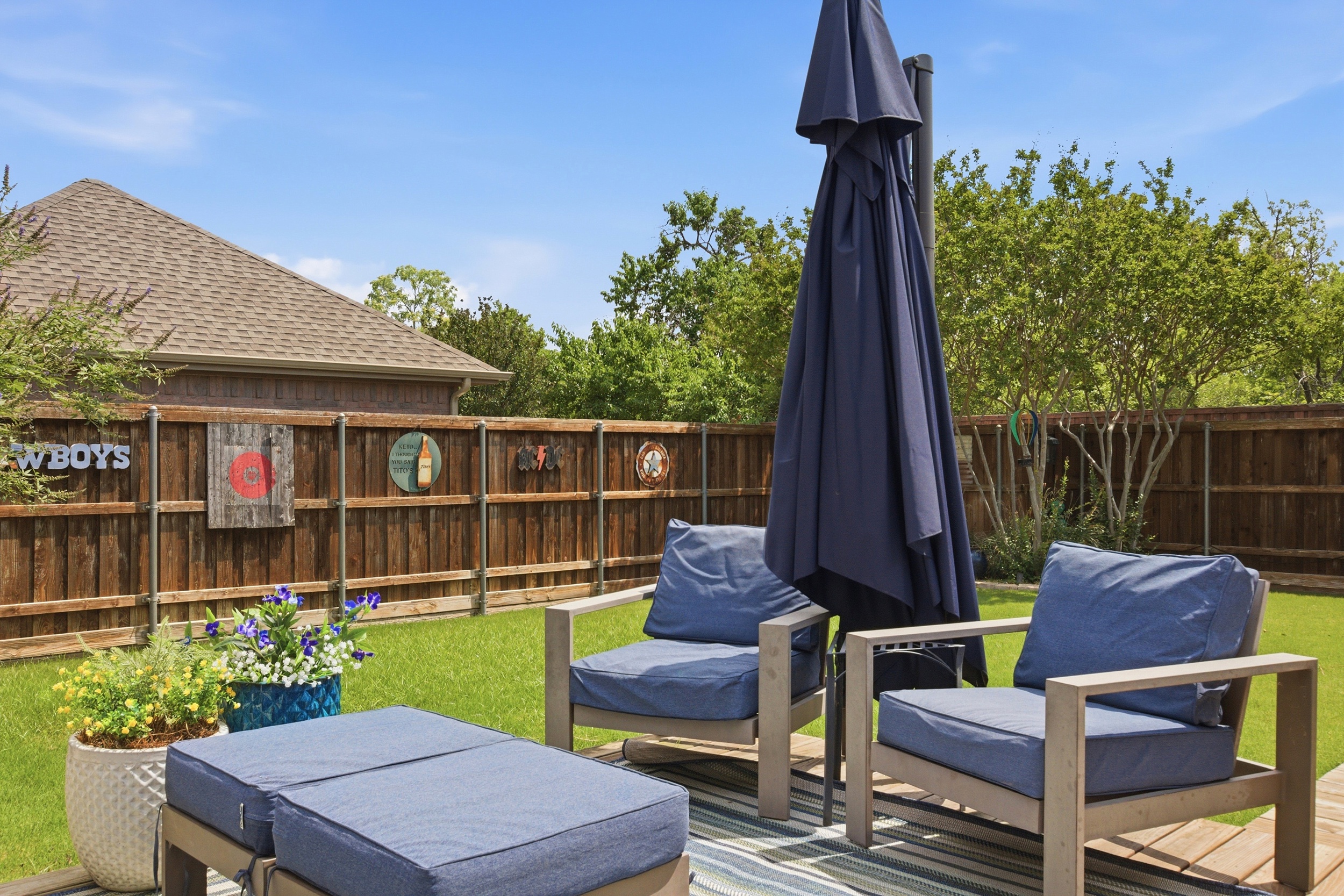 Outdoor lounge area in the Backyard with cushioned seating, side tables, and a navy shade umbrella surrounded by mature landscaping. Wall-mounted Outdoor Fan for added comfort.
