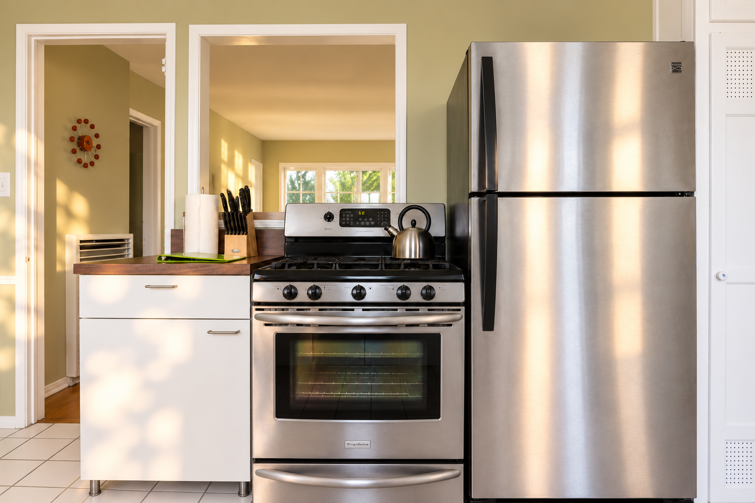The kitchen cooking area pairs a stainless-steel gas range and oven with a full-size refrigerator. White cabinetry and wood countertops frame the appliances, while the open doorway offers a view through to the sunlit living room.