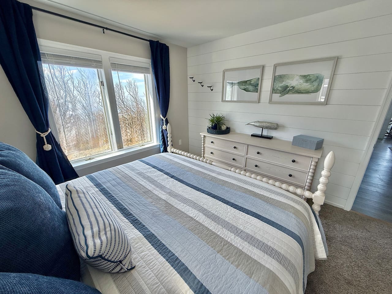 Enjoy the coastal-themed bedroom features a bed with blue-striped bedding, deep blue curtains, a light-toned dresser, and nautical wall art, creating a tranquil and well-coordinated seaside retreat.