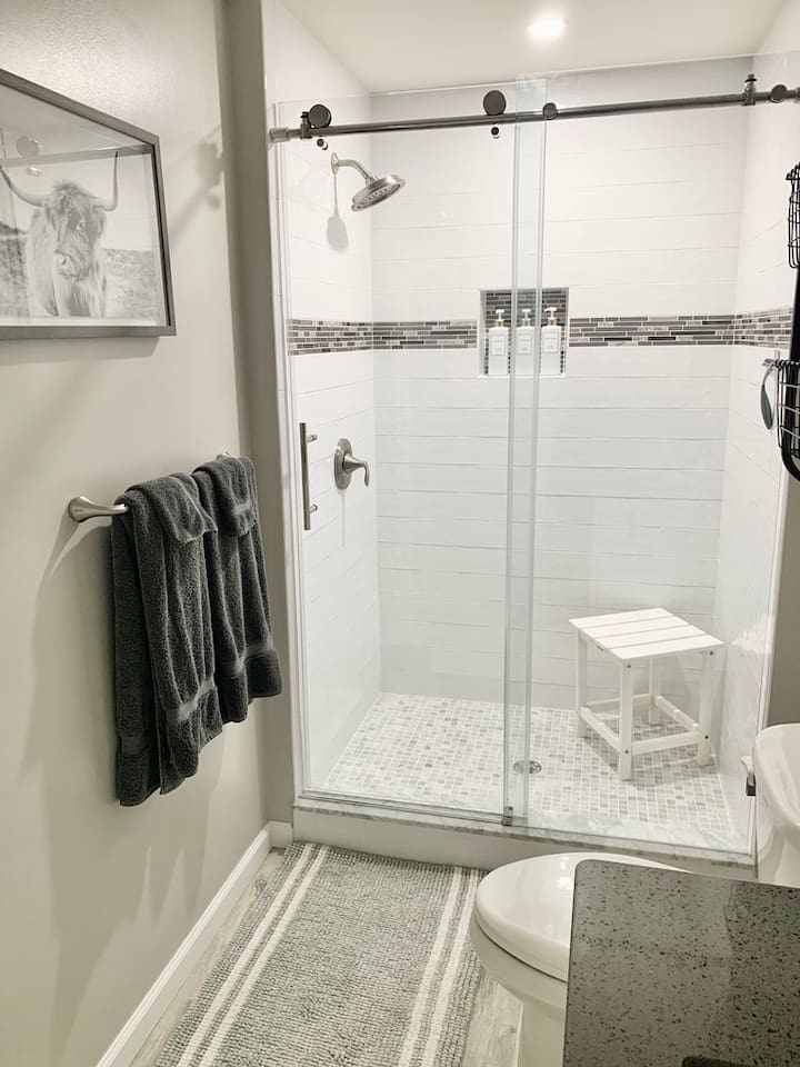 Refresh in our clean, updated bathroom featuring a spacious walk-in glass shower with sleek white subway tile and a decorative mosaic border. The shower is equipped with a convenient seat and plush charcoal towels for your comfort.