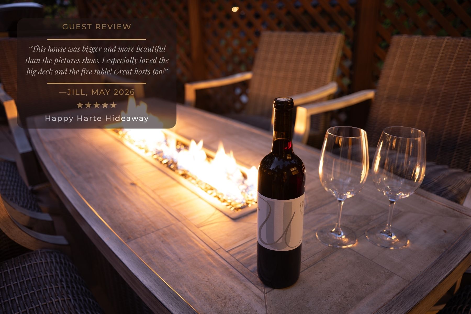 This outdoor space is a perfect evening setting, featuring a rustic wooden table with an integrated fire pit, accompanied by a bottle of wine and glasses.