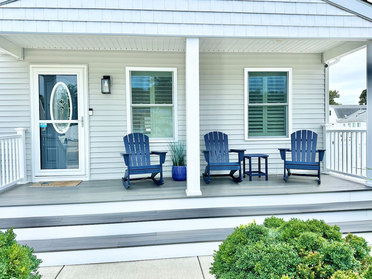 This welcoming front porch features three blue Adirondack rocking chairs and a matching side table, perfect for morning coffee. The grey composite decking and white railings provide a clean, modern look to this charming coastal entryway.