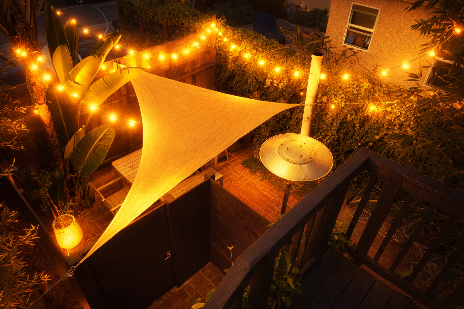 Private patio after dark, framed by lush planting and warm string lights. A triangular shade sail stretches above the timber deck, creating an intimate outdoor setting for a relaxed evening at The Venice Beachwood 2.