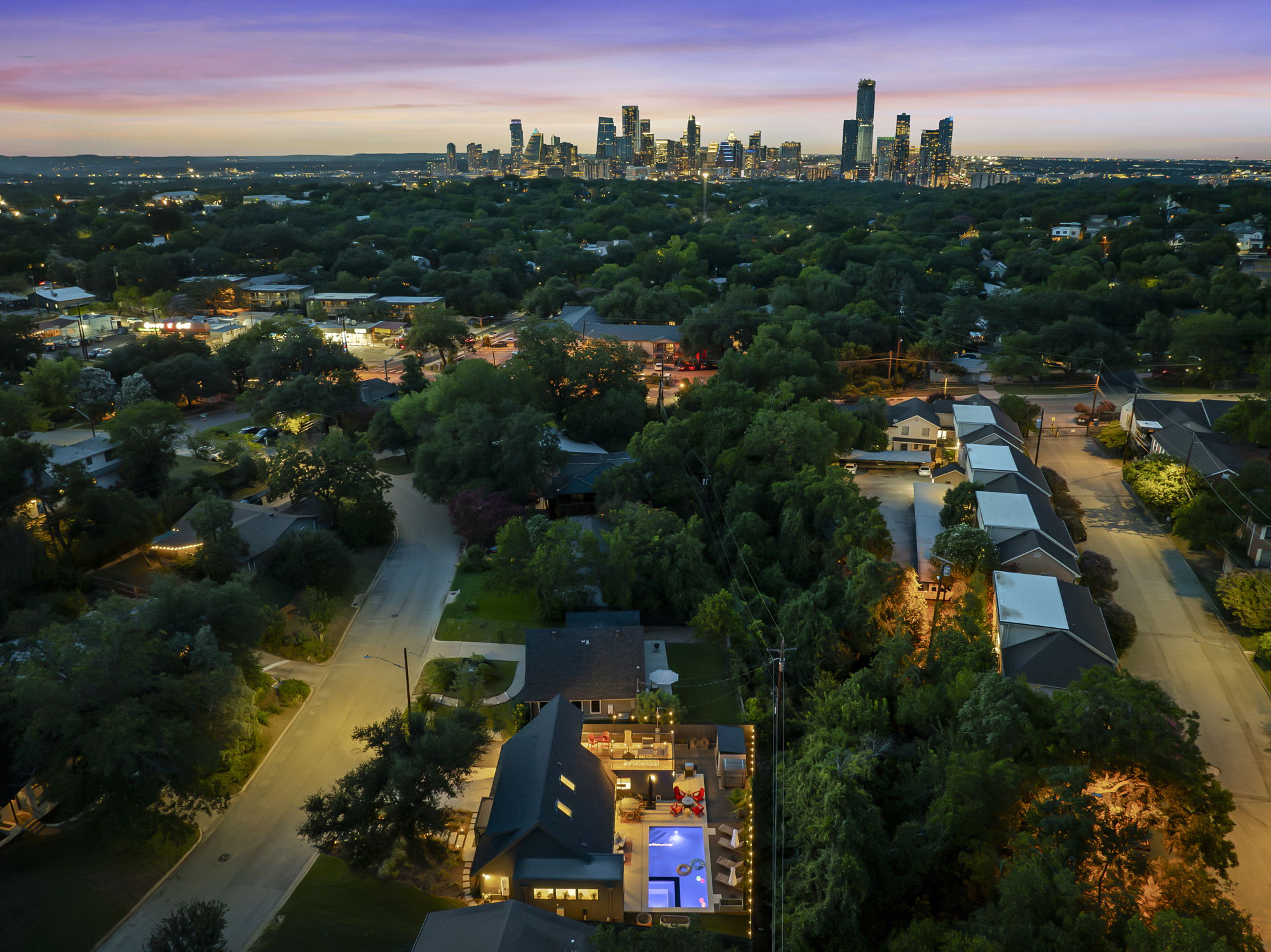 An aerial view of a modern home with a swimming pool and illuminated deck, nestled in a lush, tree-filled neighborhood with the vibrant city skyline visible in the distance.
