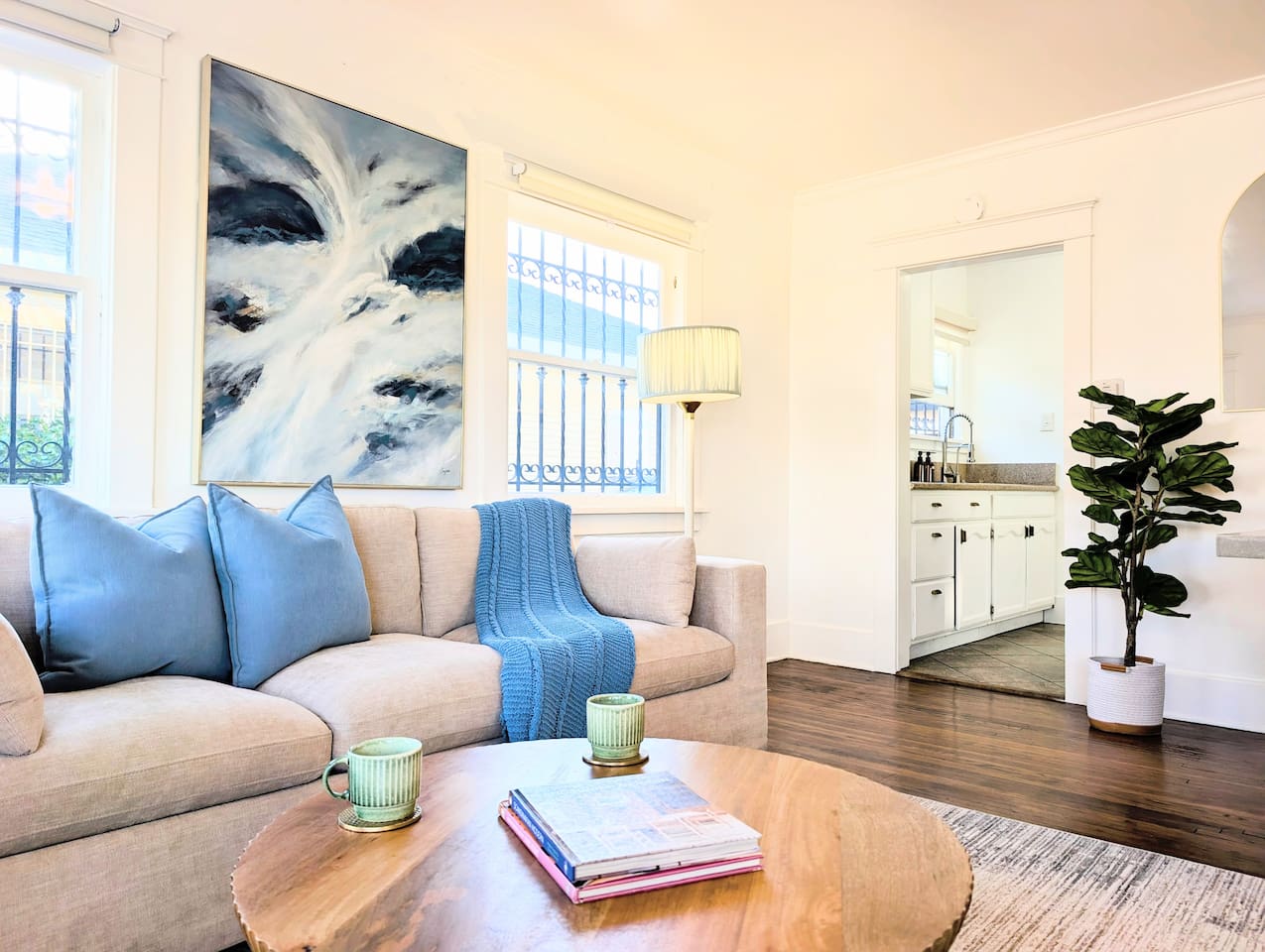 This bright, cozy living roomradiates an airy, coastal charm. Natural light floods the space, illuminating the warm hardwood floors and a plush, inviting beige sofa layered with vibrant blue accents. Framed by clean white walls, an abstract ocean painting 