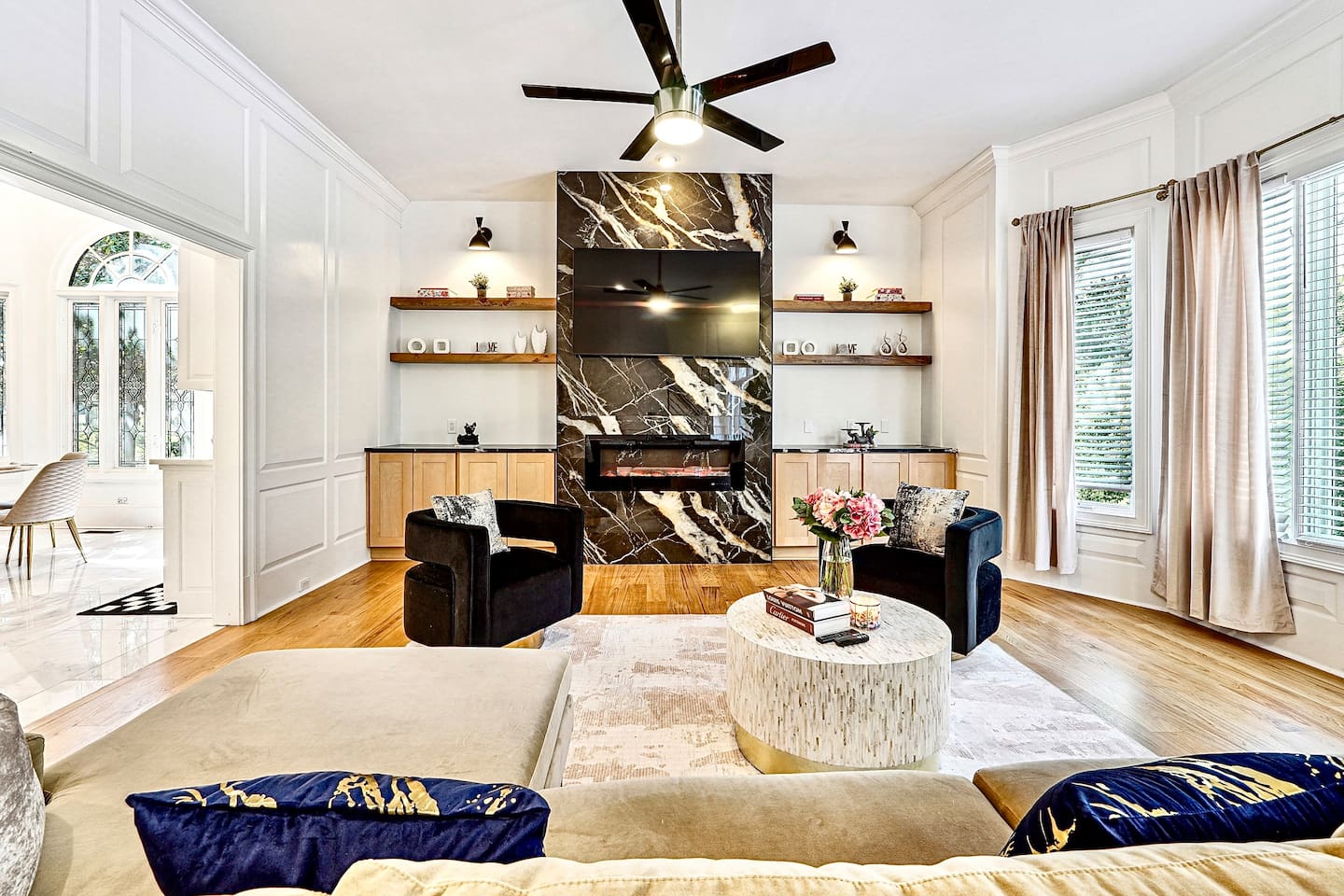 Situated next to the kitchen, this main-floor living room offers a warm, upscale ambiance with a brown L-shaped sectional, two black accent chairs, gold and black design accents, a smart TV, and a fireplace, perfect for entertaining or unwinding.