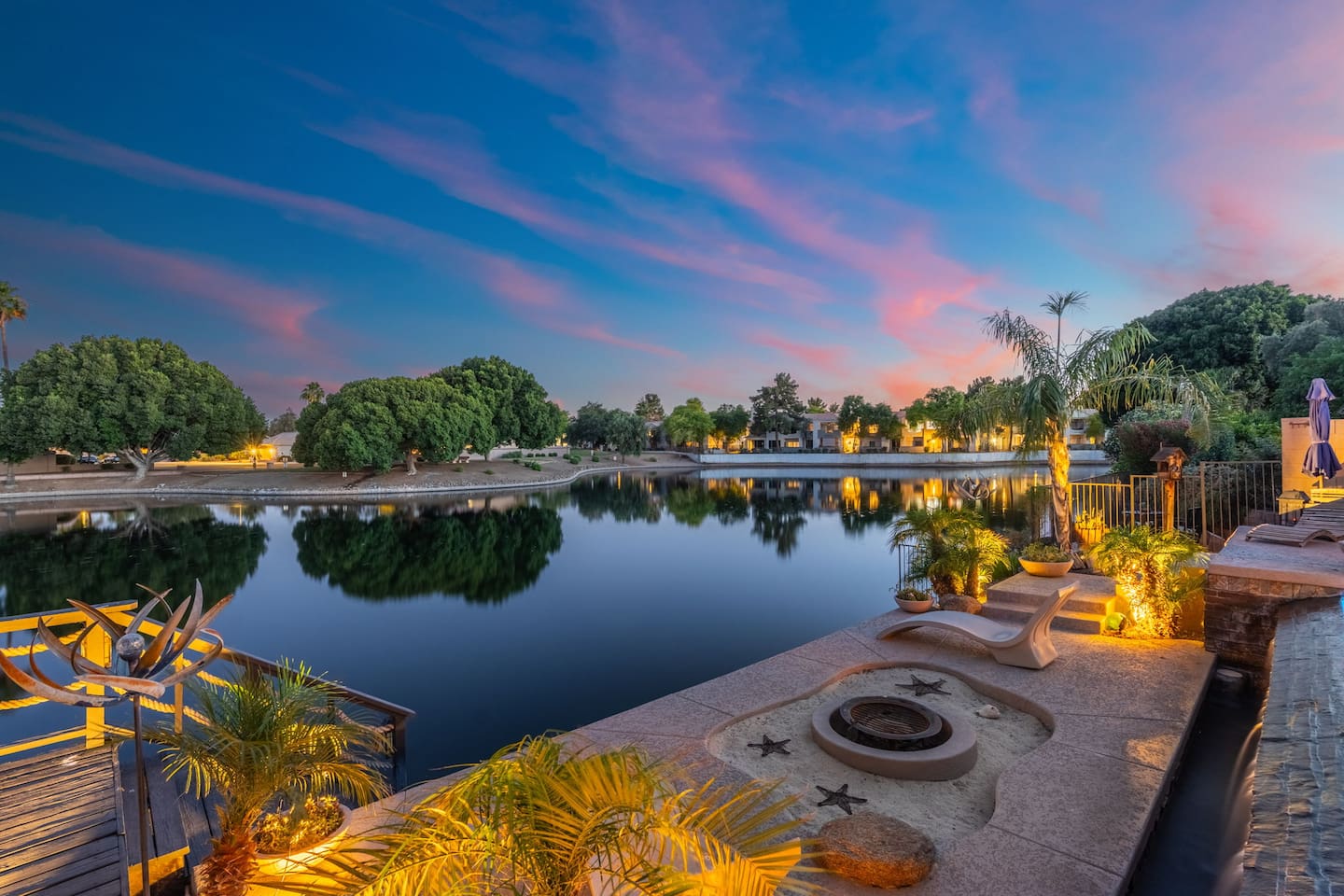The lakeside fire pit glows under a breathtaking sunset sky — gather here after dark to relax, connect, and end the day in the most beautiful way possible.