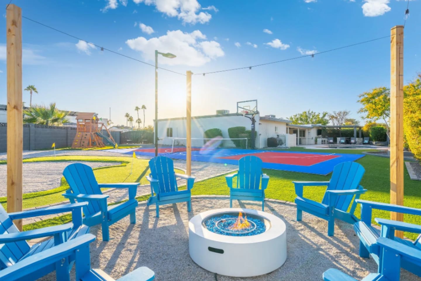 Blue Adirondack chairs ring the fire pit with a full view of the sport court and play structure just beyond, everything the family needs in one glance.
