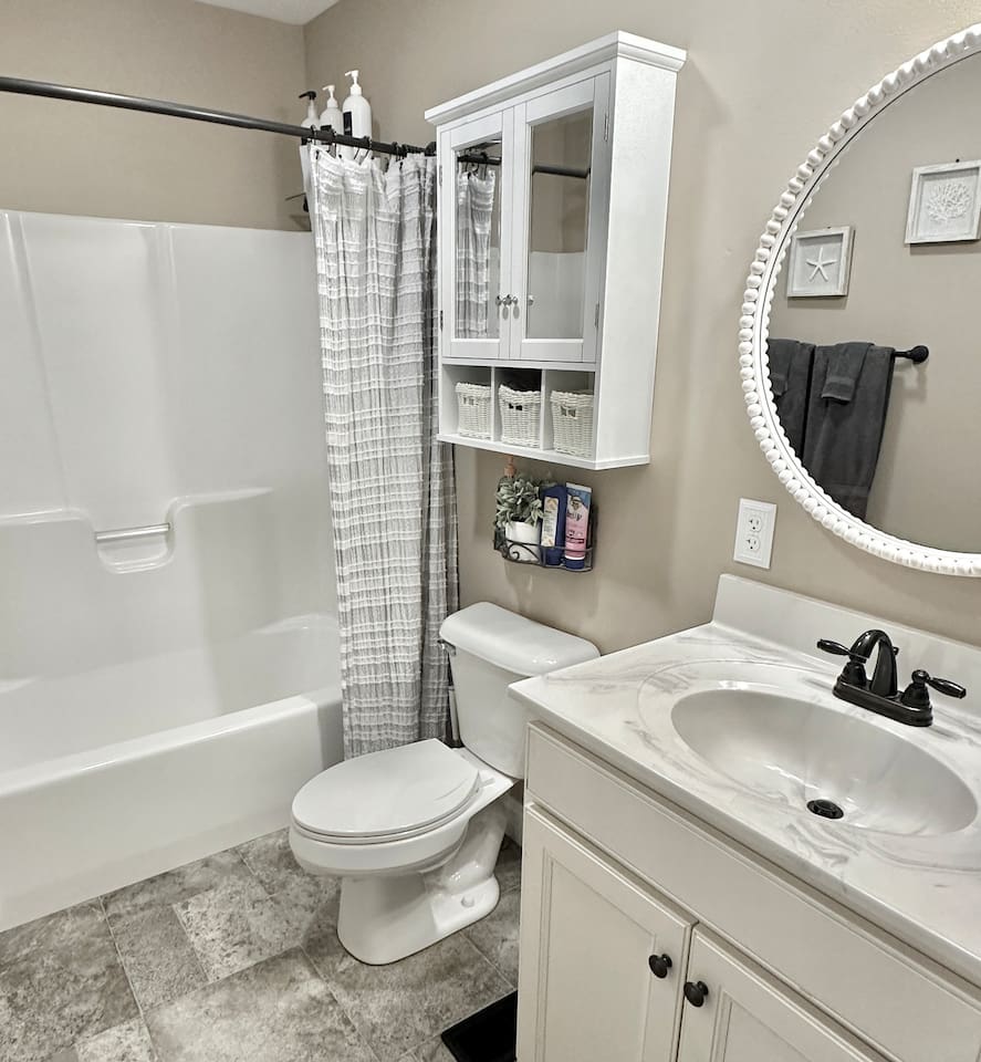 This bathroom includes a white bathtub and shower combo with a textured grey curtain. A white wall cabinet with glass doors and storage baskets hangs above the toilet, while a large beaded round mirror sits above the vanity.