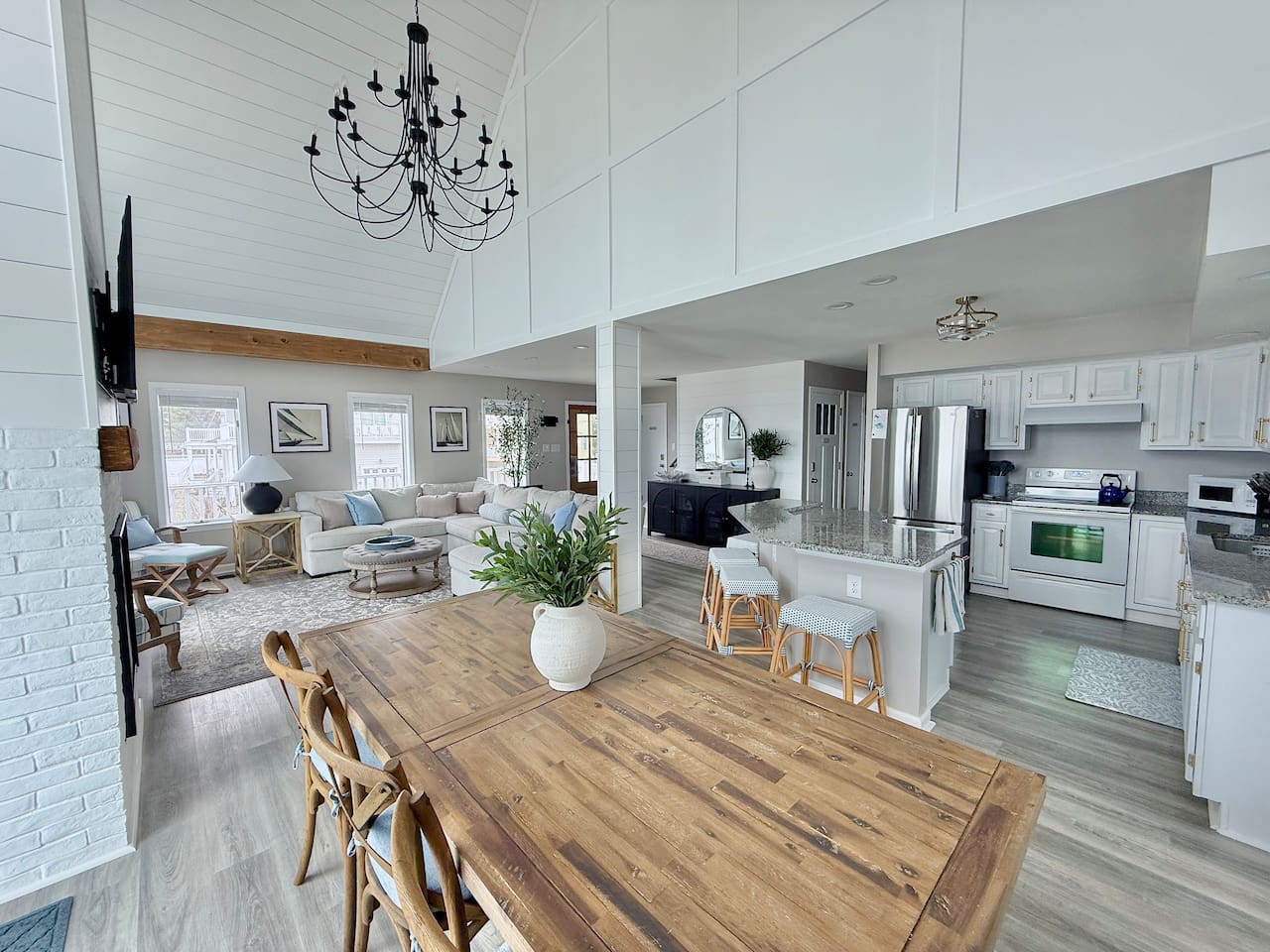 Enjoy an open-concept living space highlights a dining area with a wood table, a cozy lounge with a sofa, and a modern kitchen. A vaulted ceiling and chandelier create an airy, inviting atmosphere throughout the home.