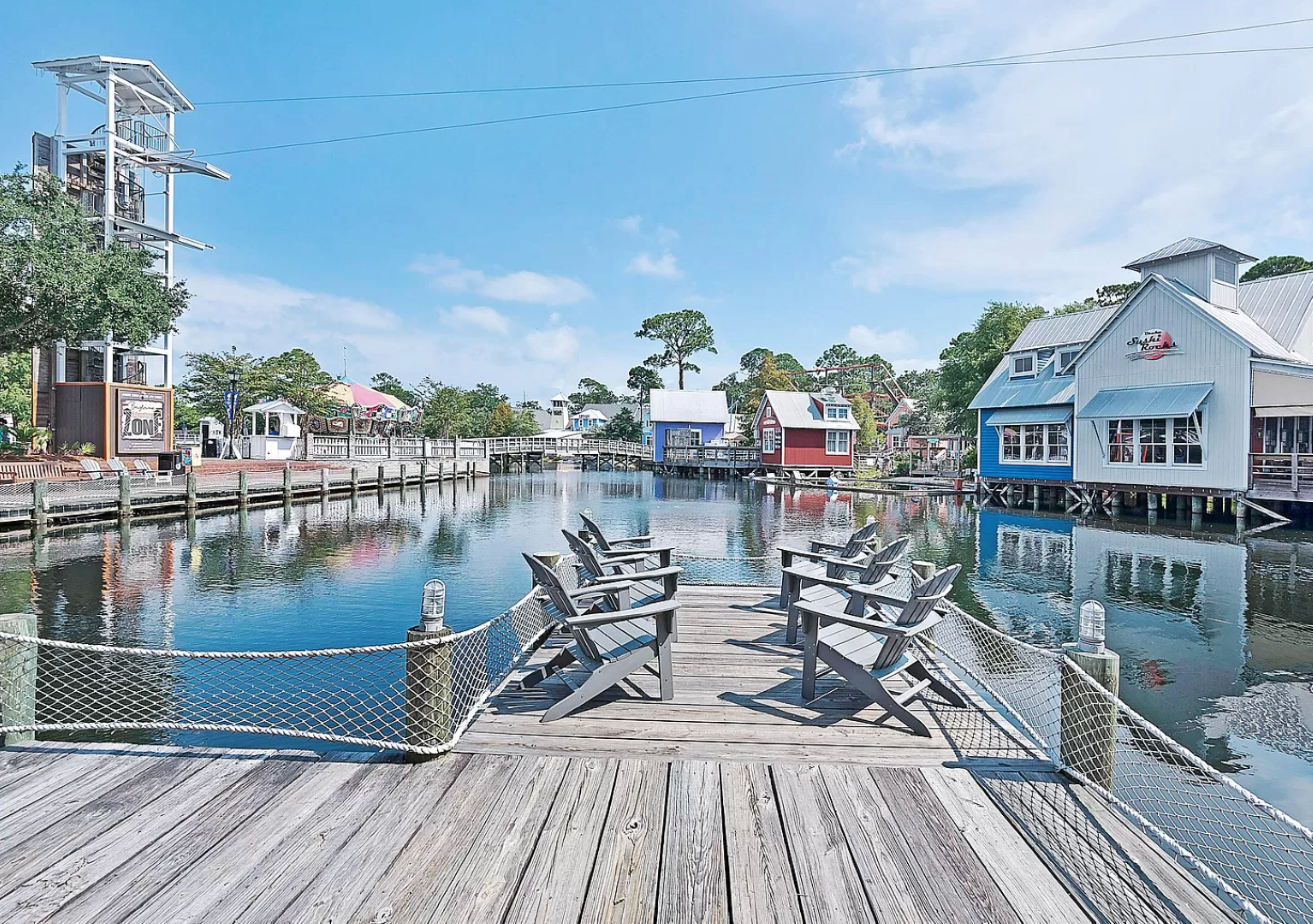Picturesque harbor landscape at The Village of Baytowne Wharf. A wooden boardwalk stretches over calm waters, flanked by colorful waterfront buildings and landscaped pathways, creating a charming seaside atmosphere.