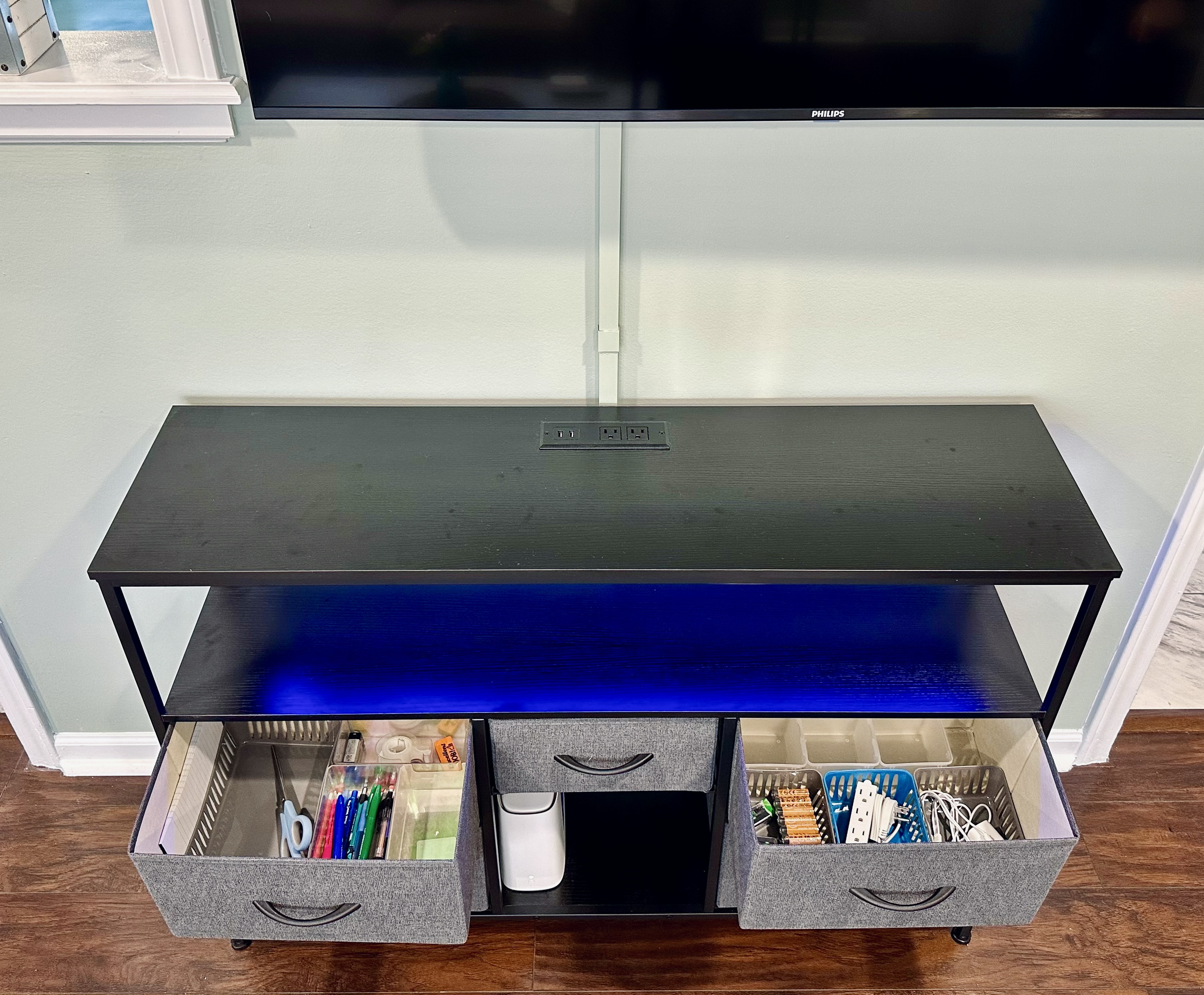 Living room TV console with deep storage drawers stocked for everyday convenience. Includes office and household essentials such as notepads, pens, scissors, batteries, charging cables, and an extension cord.