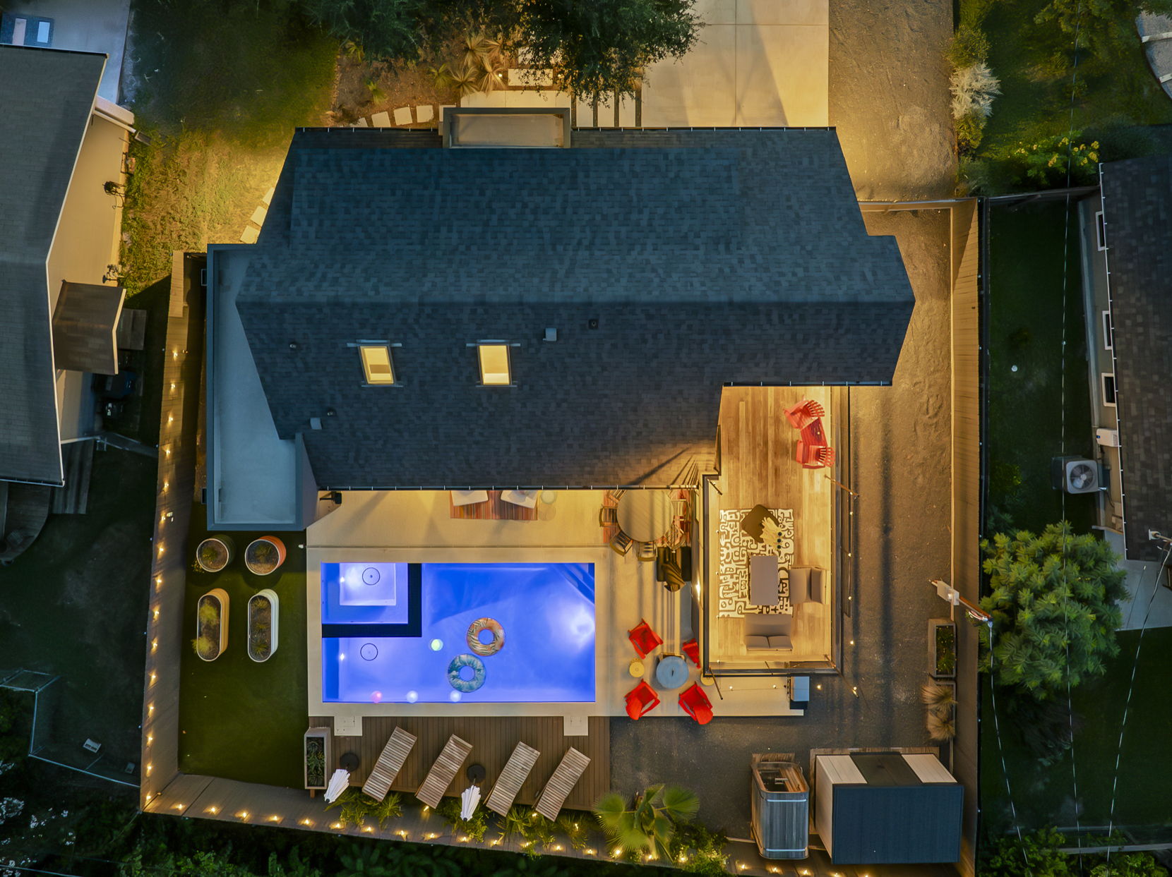 An aerial nighttime view of a modern backyard oasis, featuring a lit swimming pool with a hot tub, a spacious deck with lounge chairs and seating areas, and a lush green lawn, all illuminated to create a perfect space for outdoor entertaining.