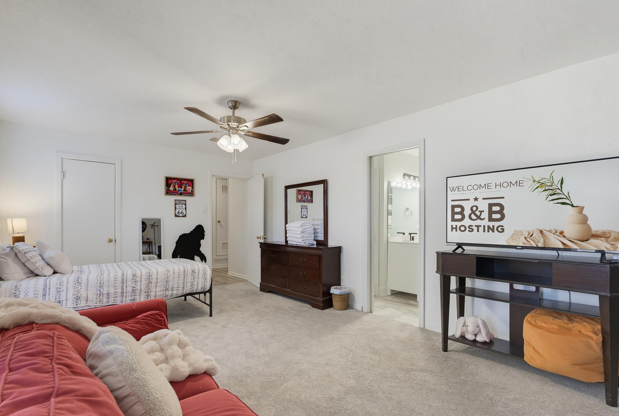 Bunk Room: An airy, open bedroom nook with a plush red sofa seating area, bunk beds and dark wood dresser topped with a mirror and folded linens. This space connects directly to a private en-suite bathroom, visible just off to the side.