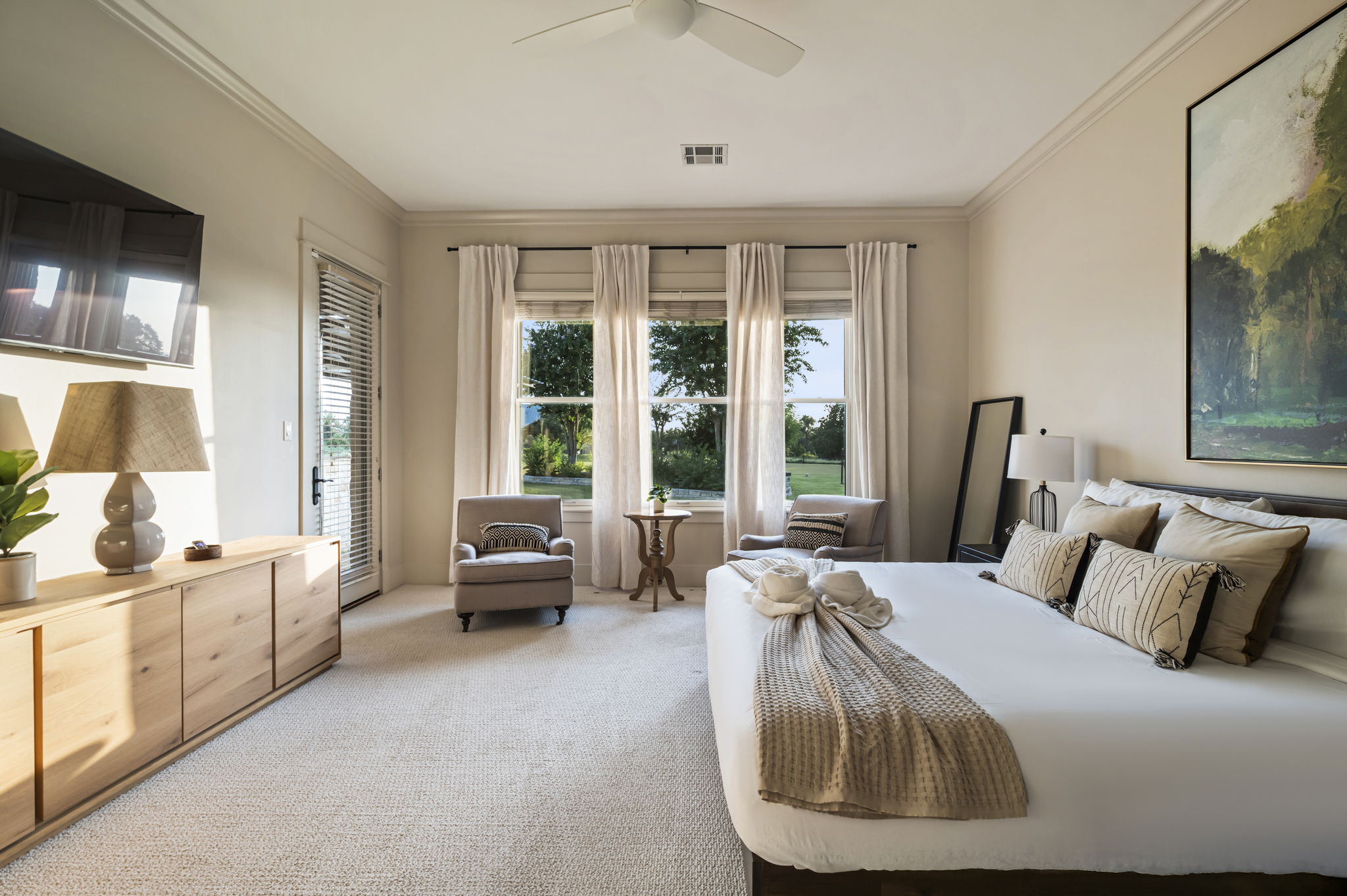 The room includes two cozy lounge chairs with a center table, a full-length mirror, two bedside lamps, and large windows that fill the space with natural light.