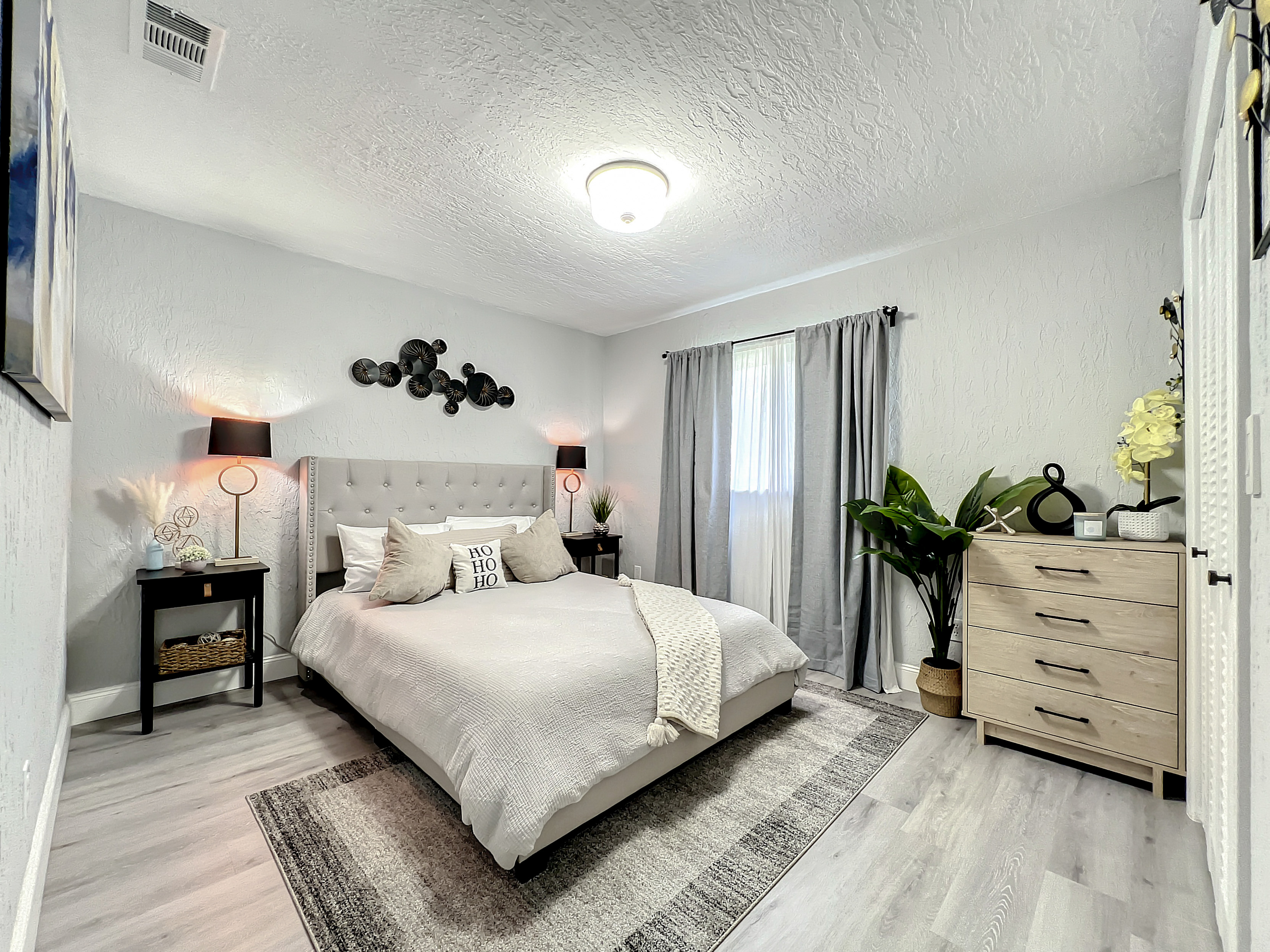 Bright bedroom with neutral decor, tufted headboard, and two bedside tables; features natural light and stylish wall art.