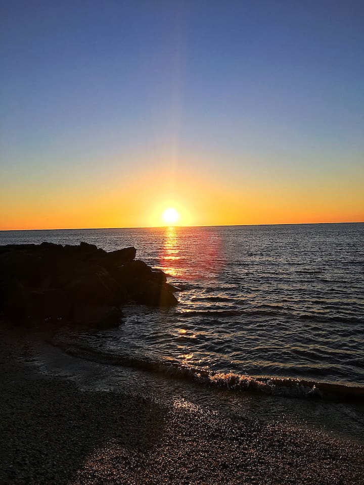 Witness stunning waterfront sunsets. The sun dips below the horizon, reflecting brilliant orange light across the rippling waves and rocky jetty for a truly magical evening view.