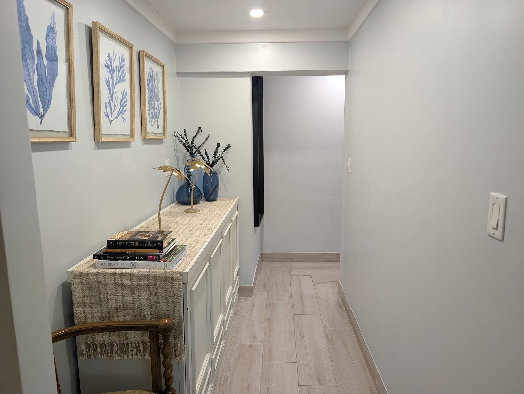 Downstairs hallway at Villa Coral featuring light wood-look tile floors, a white credenza with coastal decor books, blue glass vases, and framed botanical seaweed artwork in Dorado, Puerto Rico.
