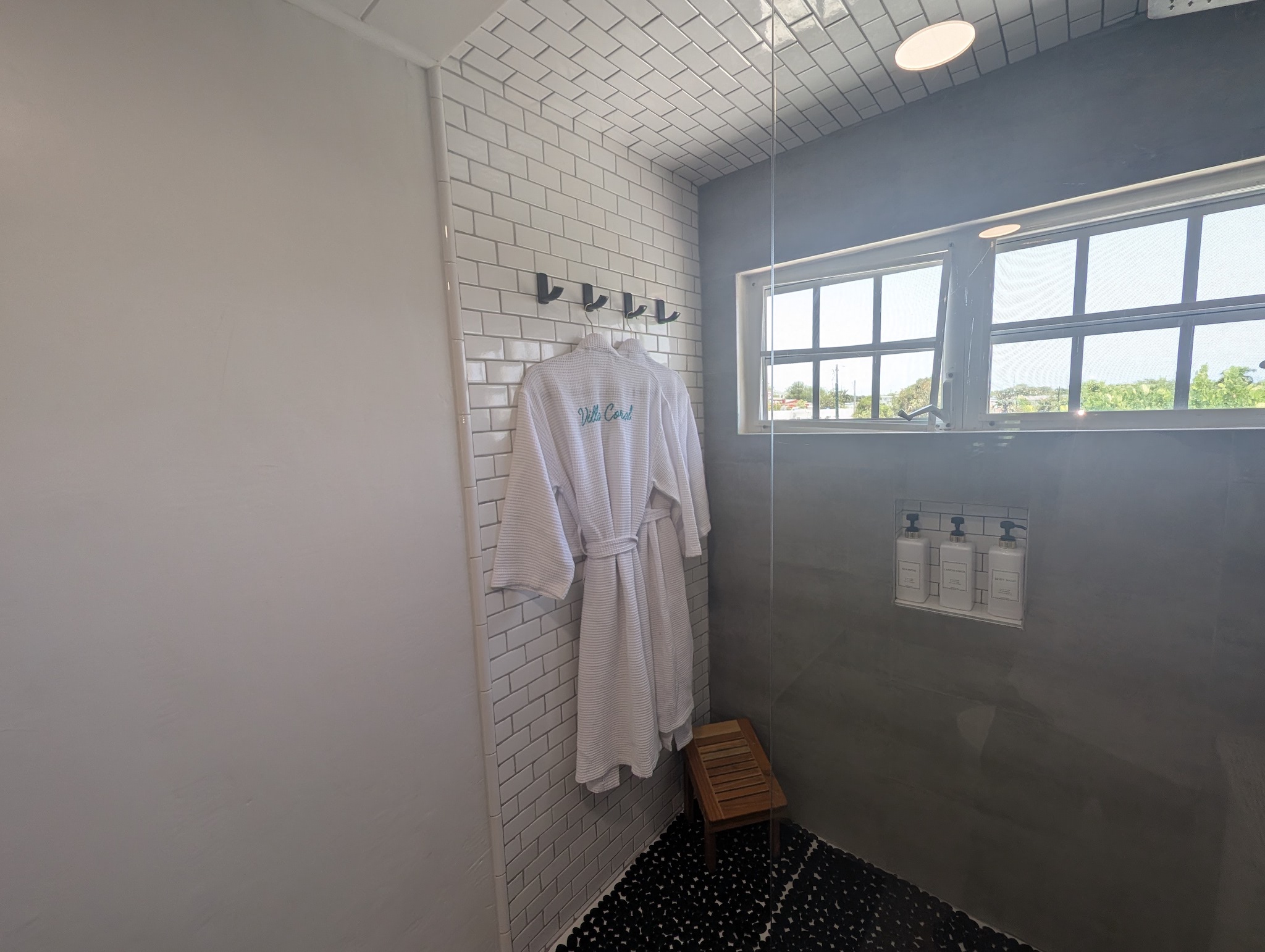 Walk-in shower at Villa Coral featuring white subway tile, large gray porcelain panels, dark pebble flooring, a teak shower stool, and embroidered luxury spa robes.
