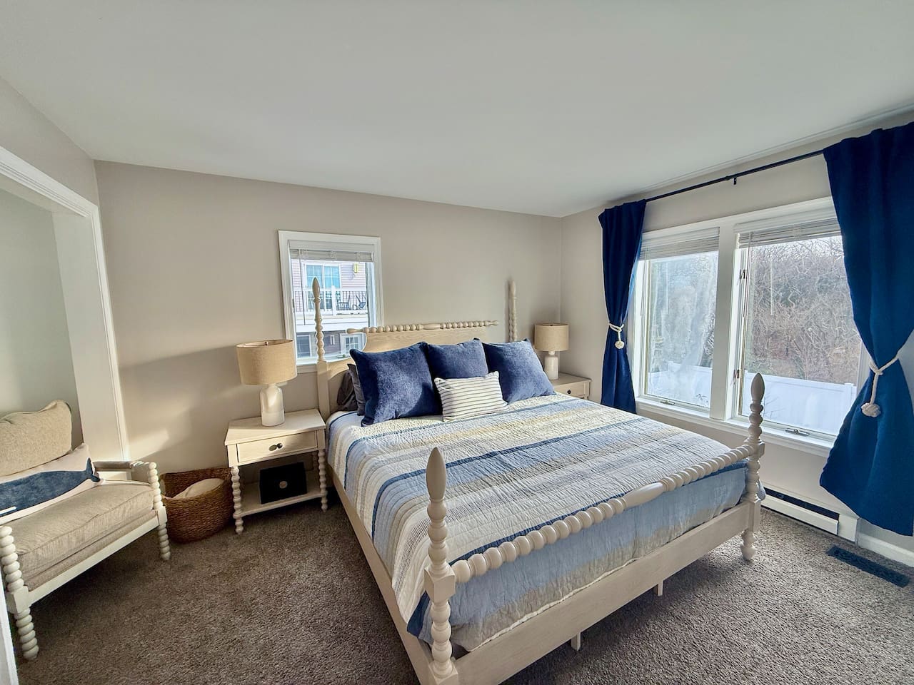This bright bedroom features water views with a white spindle bed with blue-striped bedding, a cozy armchair with a whale pillow, and deep blue curtains, creating a tranquil, well-coordinated seaside retreat for guests to relax in comfort.