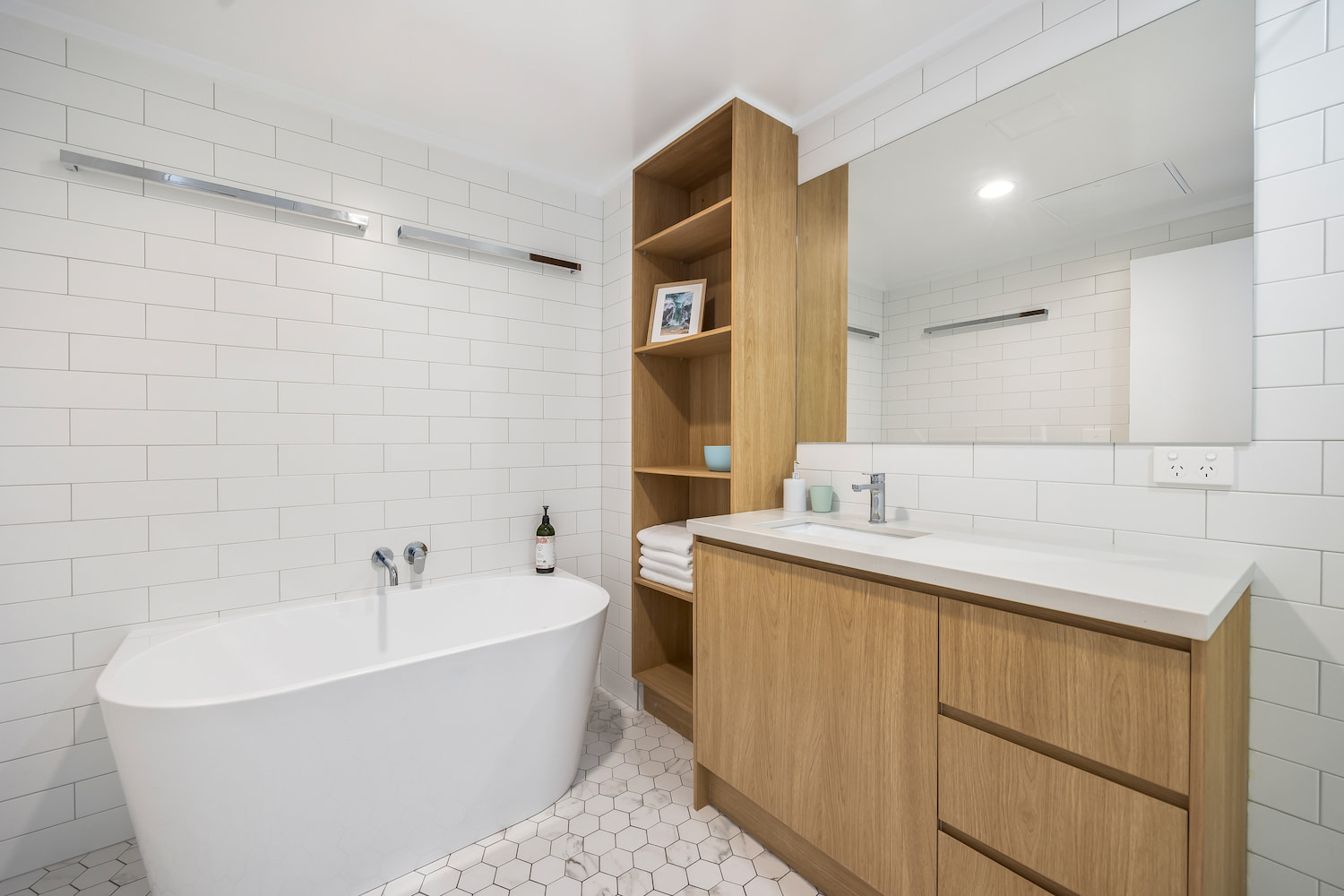 White tiled bathroom with deep bath, sink, mirror and open wood look shelves.